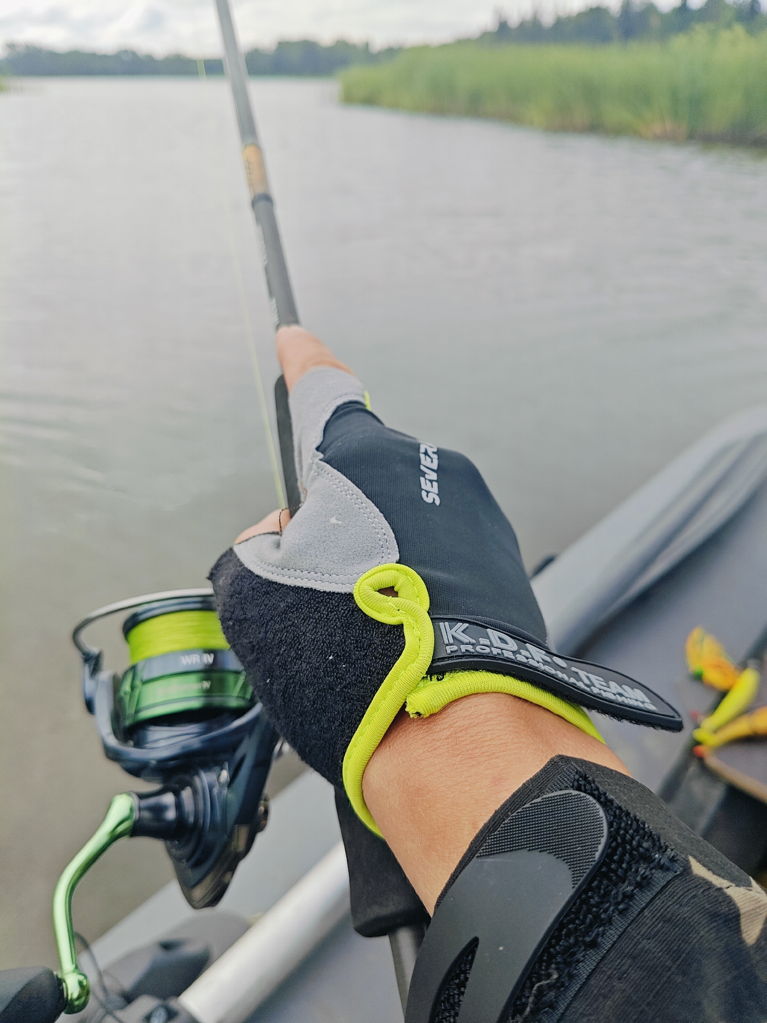 The image shows a close-up of a person's hand and arm wearing a fishing glove, holding a fishing rod. The fishing reel is visible, with bright green fishing line. The background is a body of water with a treeline and overcast sky. The setting appears to be outdoors, near a lake or river. There are no visible buildings or landmarks that would suggest a specific location within Detskii Dom (Bashenino), Russia. The focus is on the fishing equipment and the action of holding the rod.