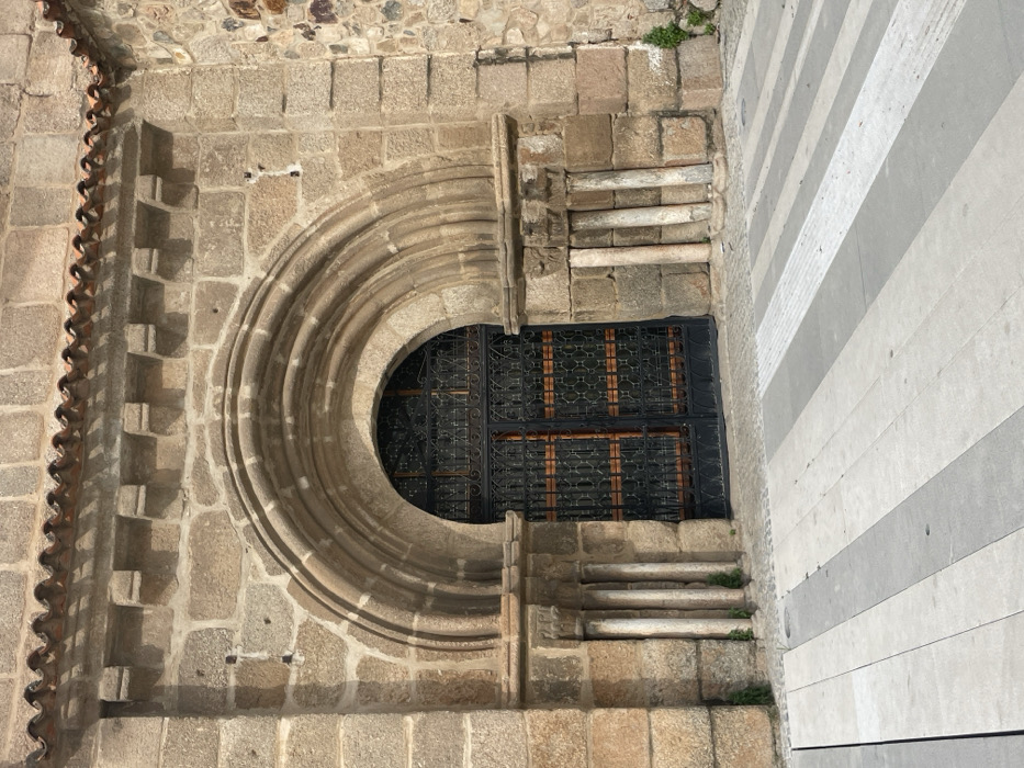 This image presents a close-up view of the exterior wall of a historic building in Mérida, Spain, bathed in bright daylight. The primary focus is an elaborate, partially visible circular window set into a robust stone wall. This stone wall is constructed from irregular, light brown and grey blocks, exhibiting a weathered texture.
The circular window, characteristic of a rose window, is framed by multiple layers of carved stone molding, creating depth and architectural detail. Within its lower half, a dark, intricate iron grill with a repeating geometric pattern secures the opening. Directly beneath this, a rectangular window, also protected by a matching dark iron grill, is visible.
To the left of the main stone wall, a decorative vertical element is present, composed of a series of terracotta tiles arranged in a wavy, scalloped pattern, suggesting an ornate architectural feature or roofline. On the far right, a contrasting modern wall with distinct horizontal stripes of alternating light and dark grey materials, possibly stone or concrete, stands adjacent to the older structure. Small tufts of green vegetation sprout from cracks in the ancient stonework. The lighting indicates a sunny day, casting subtle shadows that highlight the textures of the stone. No people or activity are depicted, and no visible text is present.