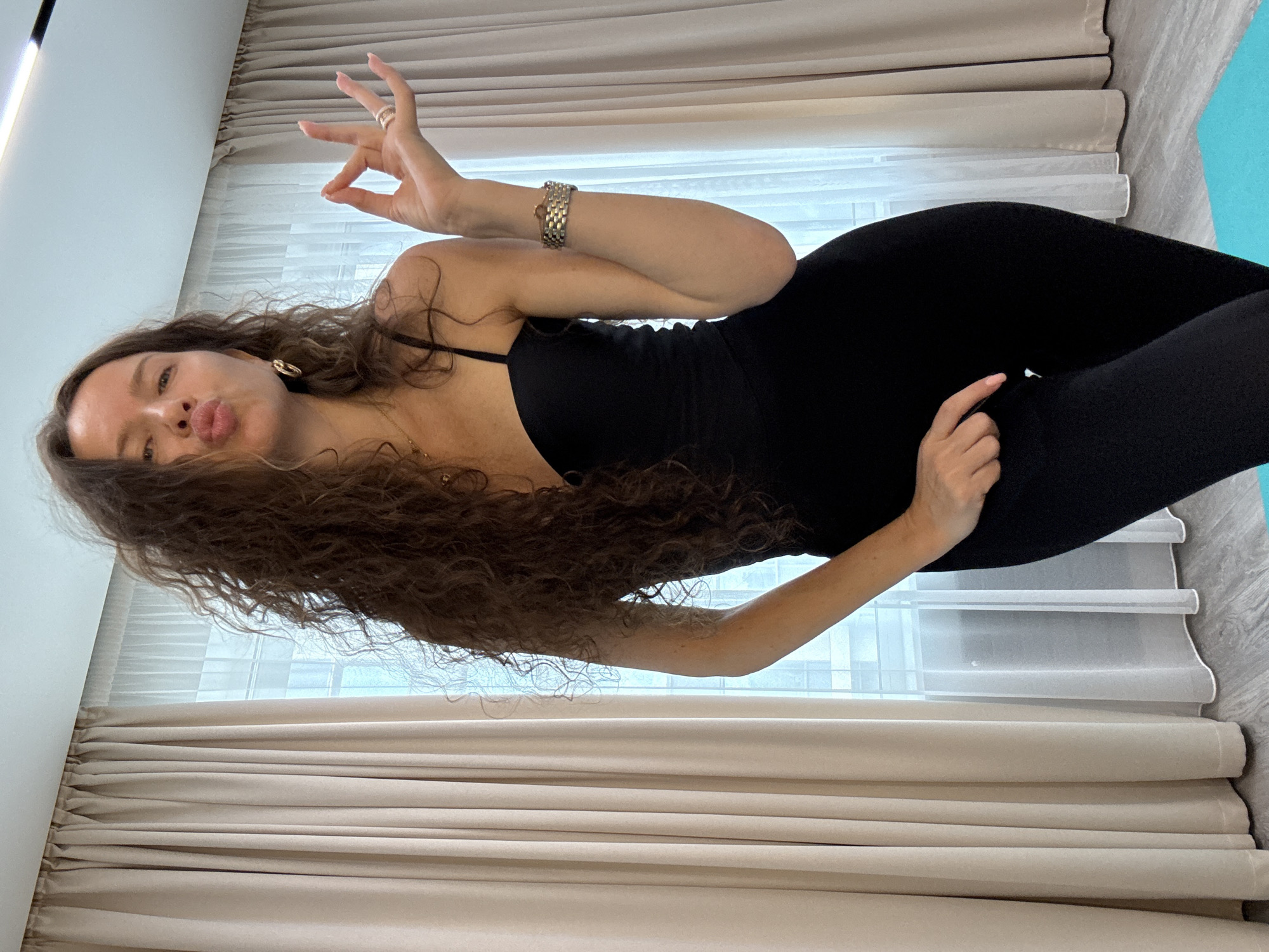 A full-body, waist-up, outdoor portrait shows a woman with long, curly brown hair. She is wearing a black, sleeveless, form-fitting jumpsuit and a gold bracelet on her right wrist. Her left hand is resting on her hip, and her right hand is raised, with her thumb and index finger forming a circle, and her other fingers extended. She is puckering her lips and looking towards the camera.
The background consists of light beige curtains drawn to the sides, revealing a window with sheer white curtains. The floor appears to be light wood, and a portion of a blue yoga mat is visible in the bottom right corner. The lighting suggests an indoor setting. There is no indication of the city of Khimki, Russia, in the image.