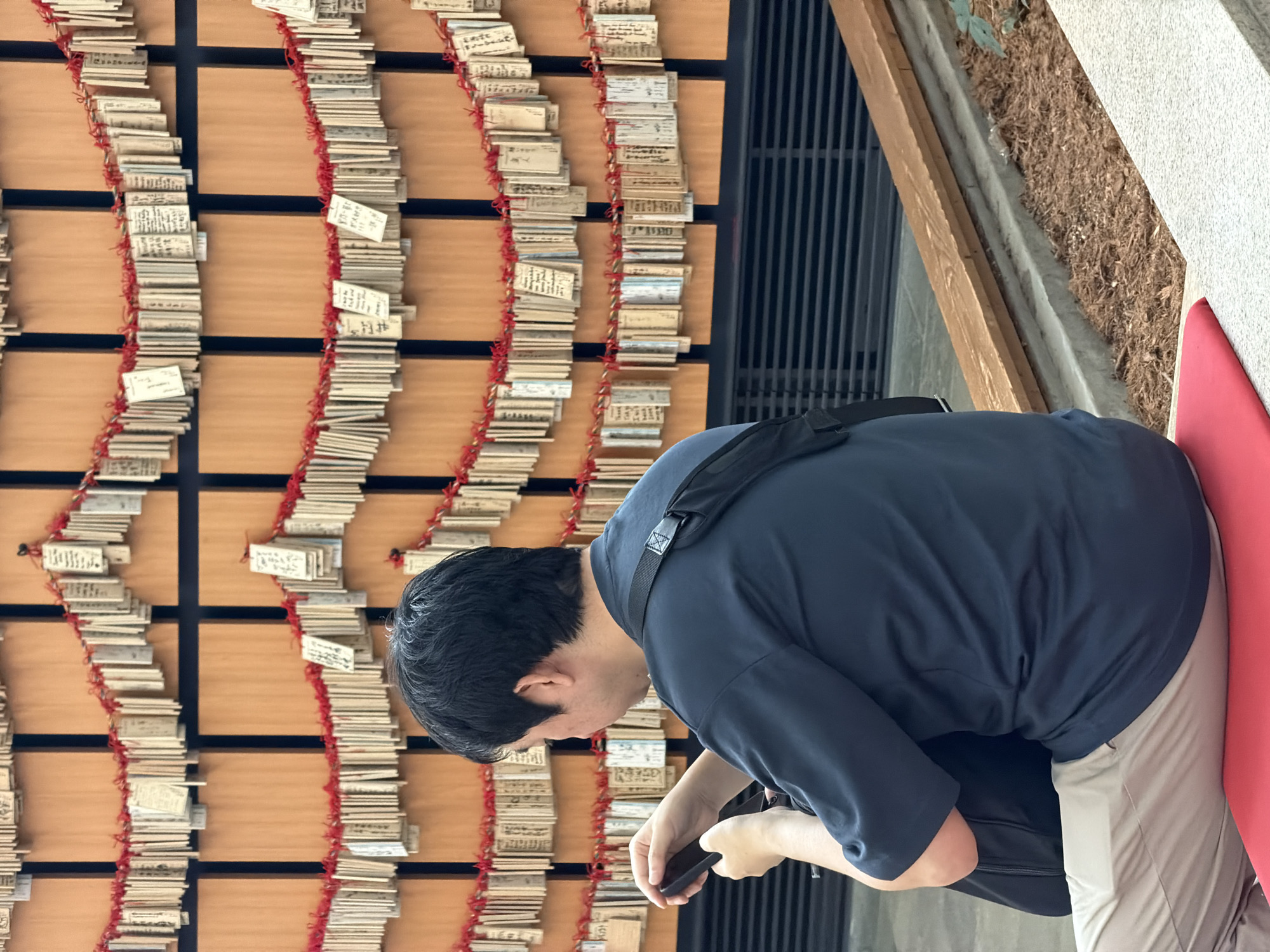 In Ota, Japan, a man is seated, facing away from the camera, looking down at his phone. He wears a dark blue short-sleeved shirt and light beige pants. A black bag is slung across his back. Behind him, a wall is covered with many small wooden plaques, each tied with red string and hanging in rows. These plaques appear to be prayer or wish tablets. The environment suggests an outdoor or semi-outdoor setting with a wooden bench and some greenery visible in the foreground.