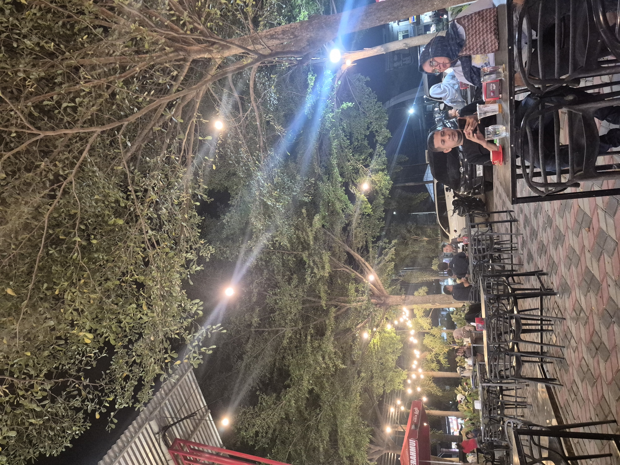The image captures an outdoor evening scene at what appears to be a casual cafe or dining area in Lhokseumawe, Indonesia. The setting is illuminated by numerous warm string lights suspended among the branches of large, leafy trees, creating a festive and inviting atmosphere against the dark night sky.
In the foreground, two individuals are seated at a metal-framed table with black chairs. A man with a beard, wearing a dark shirt, looks towards the camera while holding a mobile phone. Opposite him, another person, partially visible, wears a headscarf and glasses. Glass tumblers and a red cup are on their table. Further into the scene, more tables and black chairs are occupied by other patrons, though their faces and details are less distinct.
The ground is paved with patterned tiles in shades of red, black, and light grey. Tall, lush trees with dense foliage dominate the upper and left portions of the image, their branches crisscrossing overhead, holding the decorative lights. A glimpse of a corrugated metal roof is visible on the far left. In the background, a car with a partially visible Indonesian license plate, "BL 1647...", suggesting the Aceh province, is parked. A motorcycle or scooter is also visible nearby. On the right side, a red and white umbrella bears the text "GUDANG GARAM", an Indonesian cigarette brand. The overall impression is one of a relaxed, tropical evening gathering.