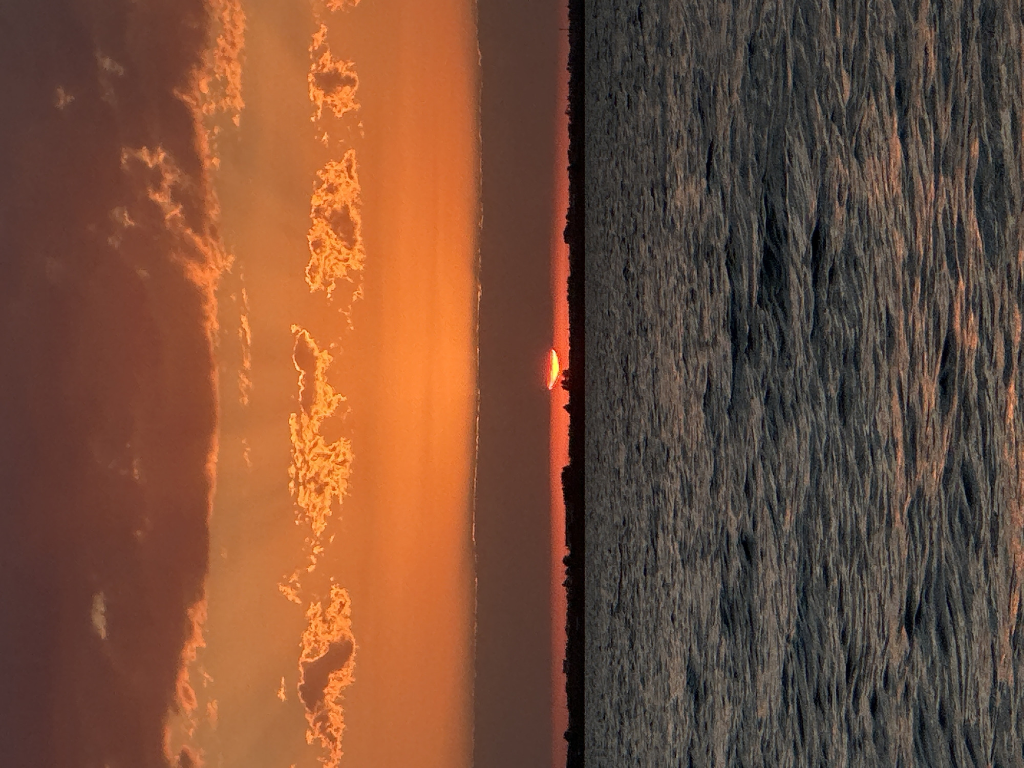 The image depicts a sunset over a body of water, likely a lake or sea, with a dark horizon line of silhouetted trees and vegetation. The sky is ablaze with vibrant orange and red hues, with scattered clouds catching the last rays of the sun. The sun itself is partially obscured by a thin layer of haze or clouds just above the horizon, appearing as a bright, glowing arc. The water in the foreground is dark and choppy, reflecting the colors of the sky. Based on the natural environment and climate, this scene could potentially be observed in Kokshetau, Kazakhstan, known for its numerous lakes and scenic landscapes during sunset. There are no people or specific identifiable structures visible in the image.