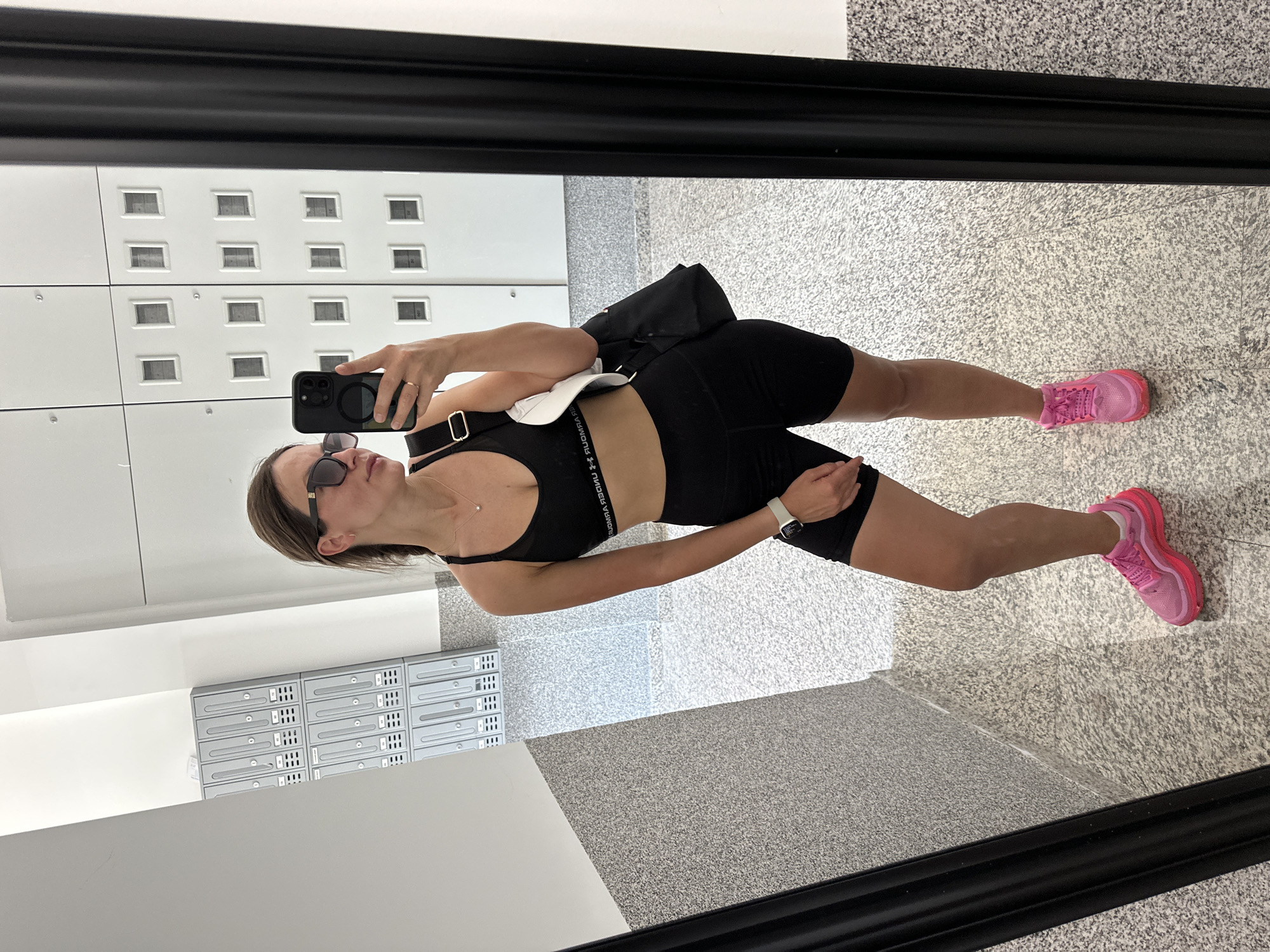 A woman is taking a mirror selfie in a hallway that appears to be in an apartment building or a similar residential complex. She is wearing a black sports bra, black athletic shorts, and bright pink sneakers. She has on sunglasses and a necklace. A black bag with a white interior is slung over her shoulder. The hallway features speckled grey flooring and walls, and on the right side, there are white lockers or storage units with small square openings. On the left side, there are gray mailboxes. The reflection shows the woman taking the photo with her phone. The image does not contain any elements that would place it specifically in Budva, Montenegro.