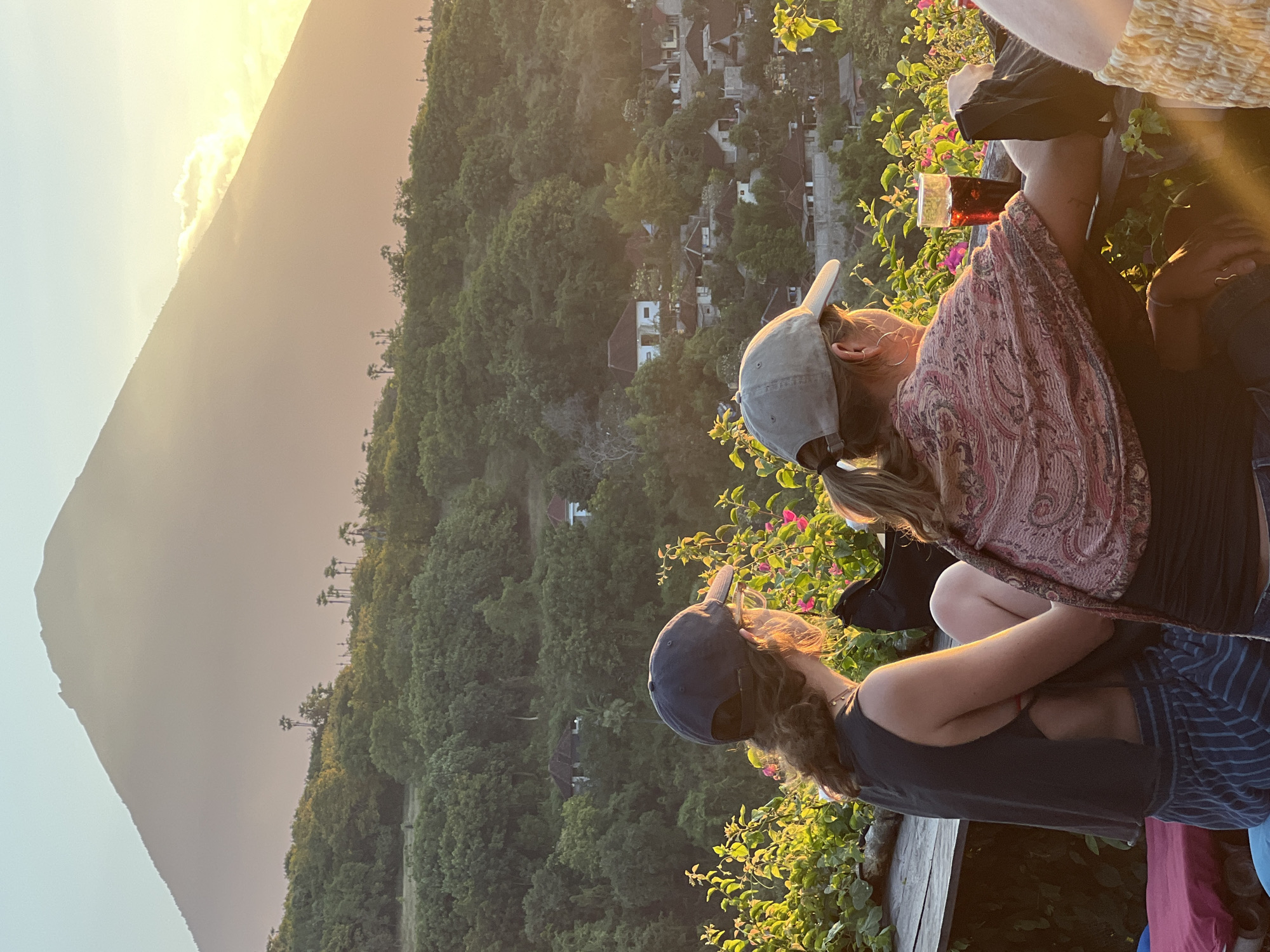 The image shows two people from the back, sitting at a wooden table in front of a view of Mount Agung in Abang, Indonesia. The person on the left is wearing a dark tank top, striped shorts, and a dark baseball cap. They have curly brown hair and are wearing glasses. The person on the right is wearing a patterned shawl over a black top and a light-colored baseball cap. They have blonde hair in a ponytail. A glass of dark red liquid is on the table in front of them, along with a black bag. The background is a lush green hillside with scattered houses and trees, leading up to the large, hazy silhouette of Mount Agung. The sun is setting, casting a warm, golden light over the scene. The atmosphere is relaxed and serene.