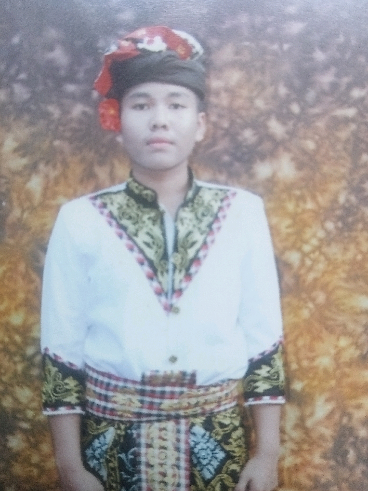 A young male stands facing the camera in what appears to be a studio portrait. He is wearing traditional clothing, including a black head covering topped with red and white fabric and red flower-like embellishments. His shirt is white with ornate gold and red embroidery around the neckline and cuffs, which are also trimmed with red and white striped fabric. A wide, patterned sash is wrapped around his waist. The background is a blurred, mottled pattern of browns and yellows, suggesting a studio backdrop. The lighting is even, illuminating the subject from the front.