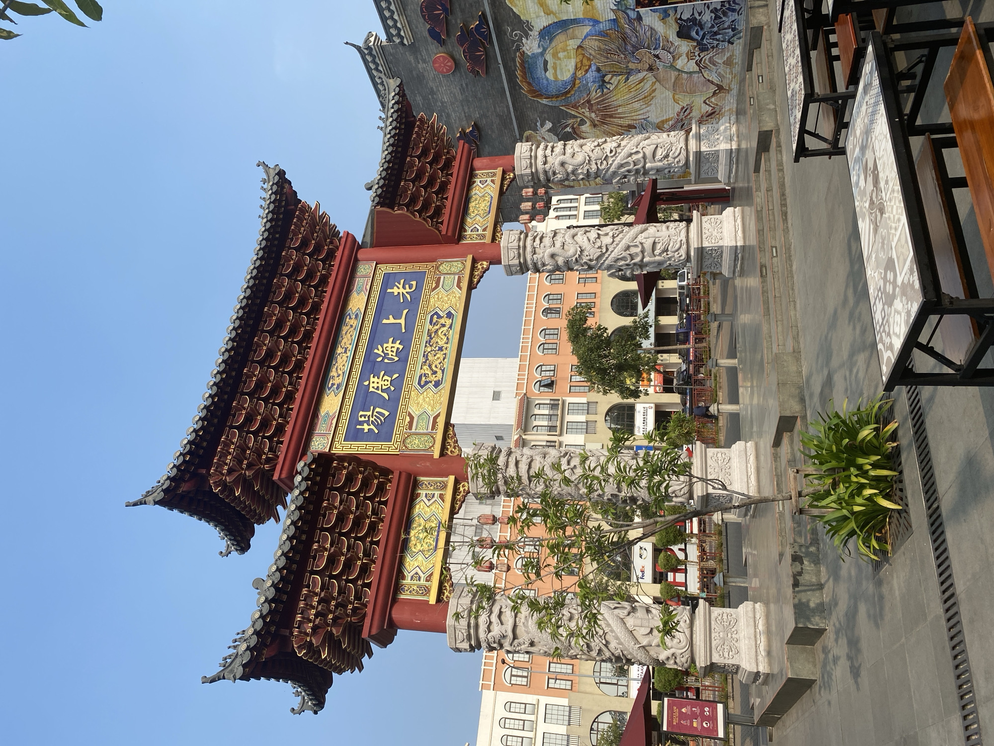 This media file depicts an ornate, traditional Chinese-style archway set against a backdrop of modern buildings. The archway is richly decorated with red and gold accents, intricate carvings on its pillars, and a large mural of a dragon above it. The pillars are carved with detailed figures, likely mythical creatures, adding to the historical and cultural feel of the structure.
The scene appears to be an outdoor pedestrian area, with tables and chairs set up, suggesting a commercial or dining space. A small tree and a lush green plant are situated near the archway, adding a touch of nature to the urban setting. The sky is clear and blue, indicating a bright, sunny day, likely in the morning or afternoon.
Behind the archway, several multi-story buildings with a variety of architectural styles are visible, some featuring arched windows and balconies. The juxtaposition of the traditional archway with the contemporary buildings suggests a location that blends different cultural influences or has been developed with a nod to its heritage.
Visible text includes "FedEx" on a sign on one of the buildings, and what appears to be a sign with "REGLASI" and some icons near the bottom of the image. The overall impression is a vibrant public space, possibly a commercial street or plaza within a larger urban environment. The setting is North Jakarta, Indonesia.