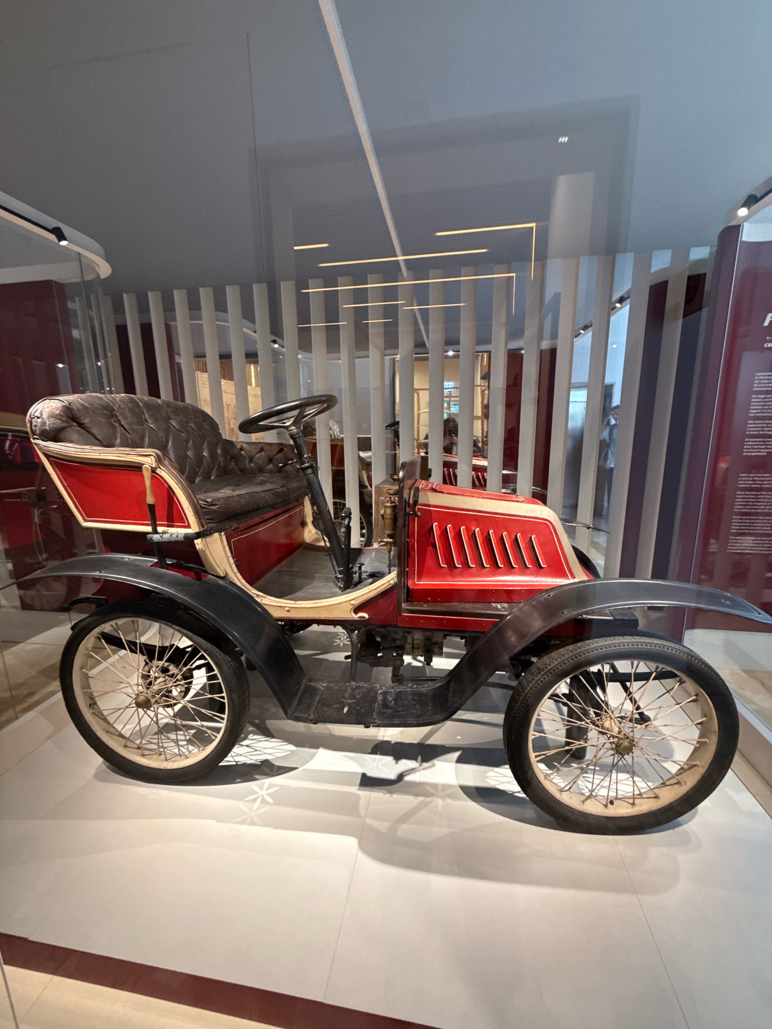 This is a full-length, side profile, eye-level shot of a vintage red and black automobile displayed indoors, likely in a museum. The car is positioned on a white tiled floor with a subtle geometric pattern. The vehicle features a brown tufted leather seat with red and cream trim, black fenders over spoked wheels, and a red hood with cream accents and vertical vent details. The background consists of vertical white slats and a red wall with some signage visible. The lighting is overhead, casting soft shadows. There are no people clearly visible in the foreground, although reflections in the glass partition behind the car suggest the presence of visitors in the background. The overall impression is that of a historical exhibit. Without further information, pinpointing the exact location within Milan, Italy is not possible based solely on the visual cues present in the image.