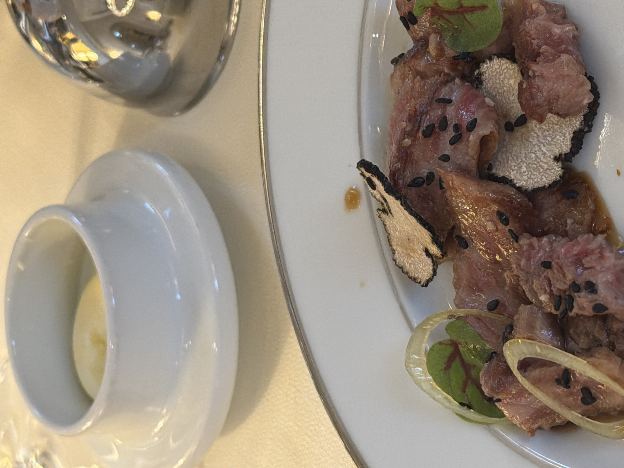 The image shows a plate of food next to a small white bowl containing a yellowish substance, possibly butter or sauce. A reflective silver object is also partially visible. The food on the plate appears to be slices of raw meat or fish topped with black sesame seeds, slices of truffle, and small green leaves. The scene likely takes place in a restaurant setting. The specific location in Belgrade, Serbia, cannot be determined from the image alone.