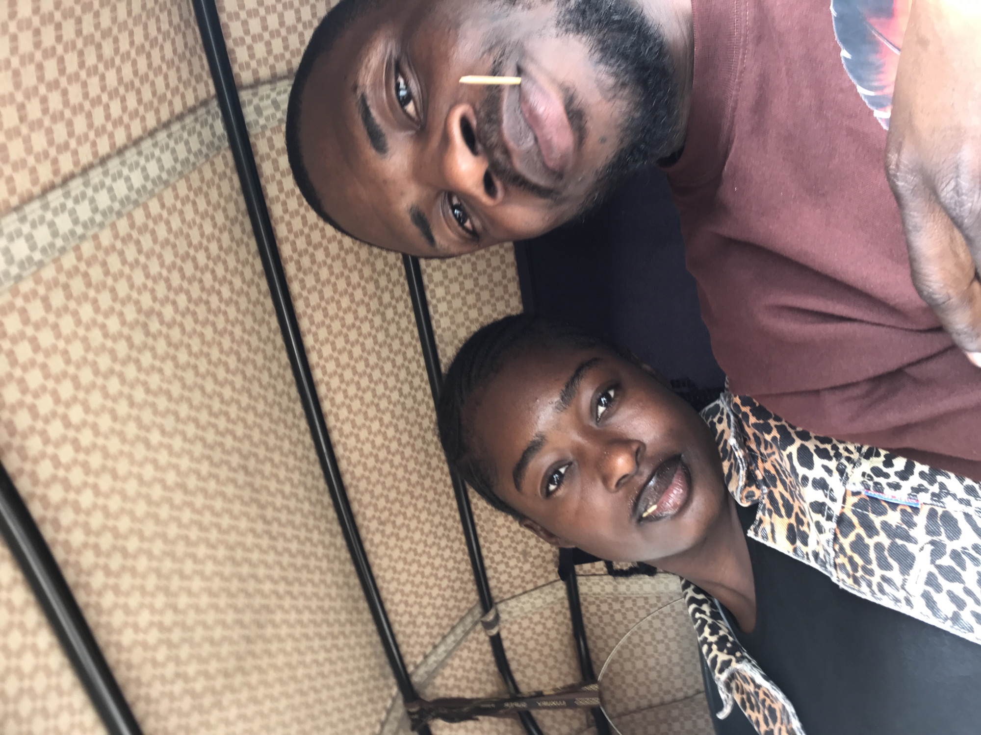 A selfie captures two individuals, a man and a woman, inside what appears to be a motorized rickshaw or tuk-tuk in New Stadium, Nigeria. The vehicle's ceiling is covered with a light brown or beige patterned fabric featuring a repeating small square or checkered design, supported by black metal bars traversing horizontally.
On the left, a woman with dark skin and braided hair looks directly at the camera with a neutral expression. She wears a black top beneath a leopard-print jacket in shades of brown and black. A light-colored, patterned bag or pouch is partially visible hanging behind her left shoulder.
To the right and in the foreground, a man with dark skin, short dark hair, and a trimmed beard holds the camera. He also looks directly forward with a neutral gaze. A wooden toothpick is distinctly visible protruding from the right side of his mouth. He wears a maroon short-sleeved t-shirt, with a partial graphic design featuring abstract red, orange, and blue elements visible on his chest. His right hand is positioned at the bottom right of the frame, holding the camera or phone used for the photograph.