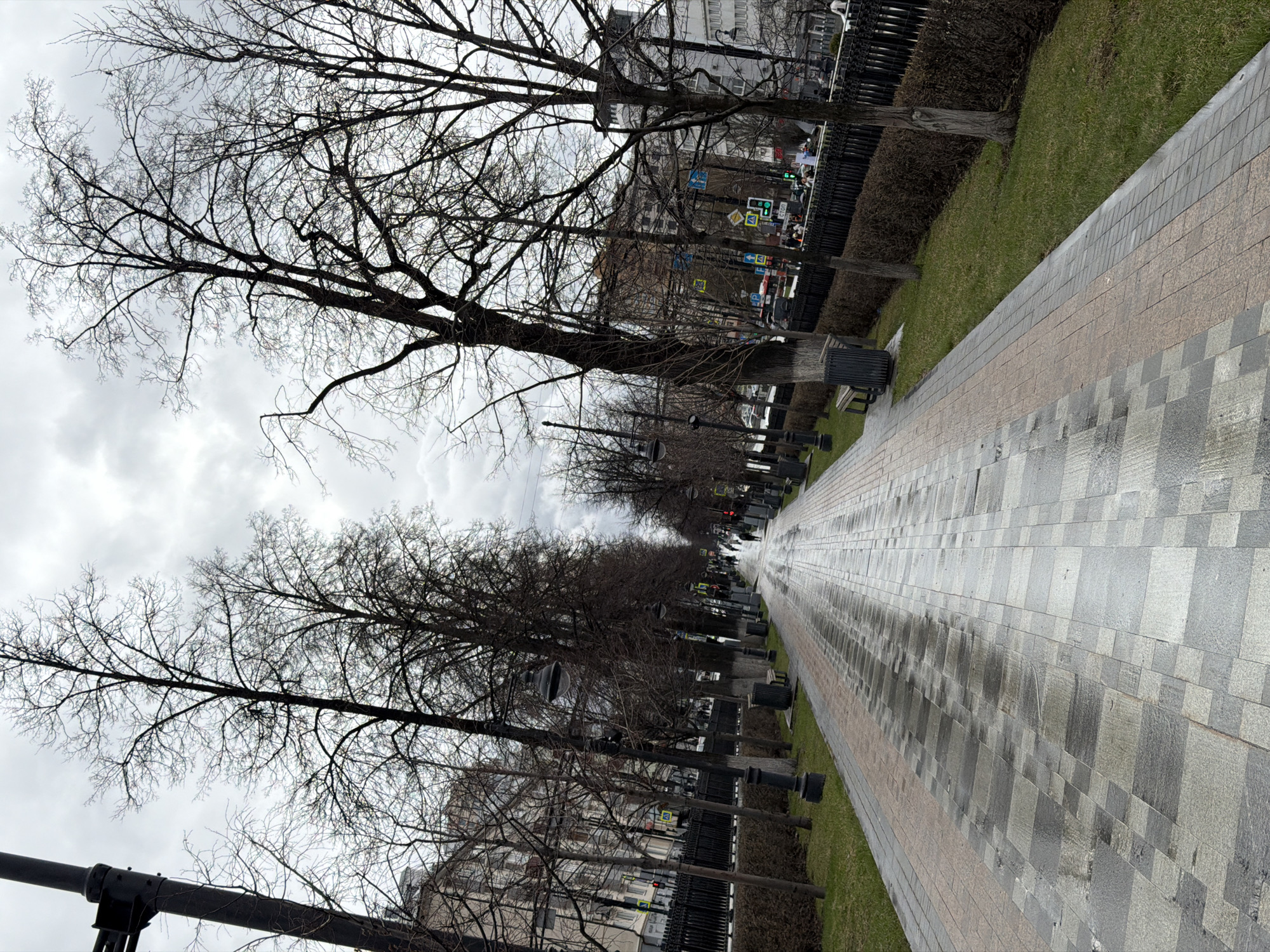 The image captures a long, straight paved walkway in Perm, Russia. The walkway, composed of grey and tan rectangular pavers, stretches into the distance, appearing wet. Along the sides of the walkway are patches of green grass, brown bushes, and rows of bare trees. Benches and ornate lampposts line the path, creating a park-like atmosphere. In the background, buildings can be seen, suggesting the walkway is within a city environment. Street signs and lights are visible, reinforcing the urban context. The sky is overcast, contributing to the muted tones of the scene.