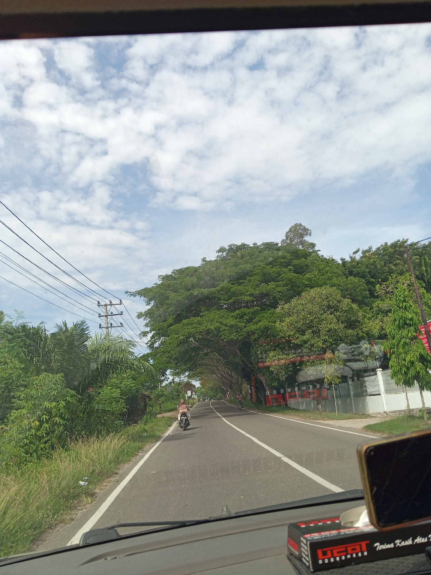 Captured from inside a vehicle, the image shows a road in Pulorungkom, Indonesia. A motorcycle is traveling along the road. Vegetation lines the roadside, including a canopy of trees, along with overhead electrical wires. On the right side of the road is a red and white building. The sky is blue with scattered clouds. A tissue box and a phone are visible on the dashboard of the vehicle.