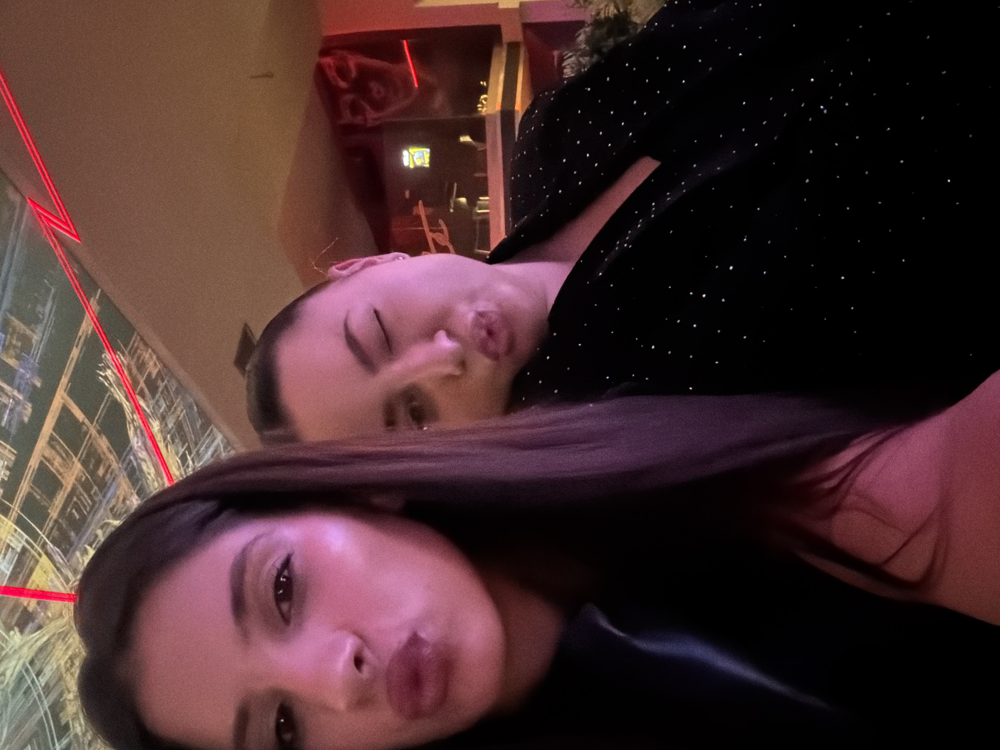 Two women are posing closely together for a selfie indoors. The woman closest to the camera has long, dark hair and is looking at the camera with a slight pout. The other woman, positioned behind and above, has her eyes closed and is also making a pouting expression. She is wearing a black top with small, shiny embellishments. In the background, there is a cityscape reflected in a window or glass panel, illuminated by red and other colored lights. The overall scene appears to be lit by a dim, red-toned light. The environment is a city, and I cannot infer that this is Sofia, Bulgaria.