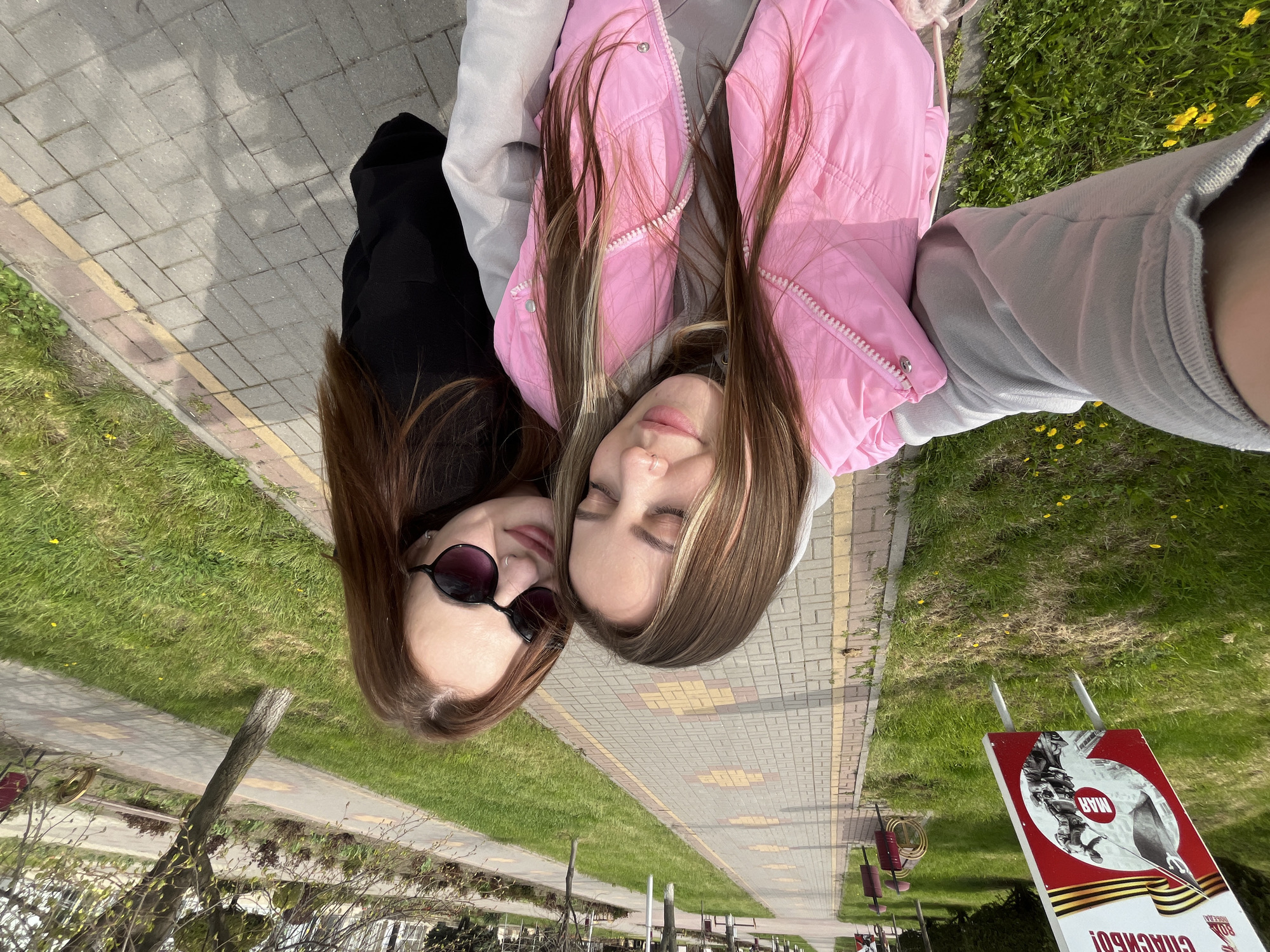 Two women are lying close together on a brick sidewalk with grass on either side. One woman wears dark sunglasses and a black top, while the other wears a pink puffer jacket and a grey hooded sweatshirt. A red and white sign in Russian stands in the grass. The sign has a military theme. The sidewalk and sign suggest the city of Novopokrovskaya, Russia, given the content of the sign. The scene appears to be a selfie taken by one of the women.