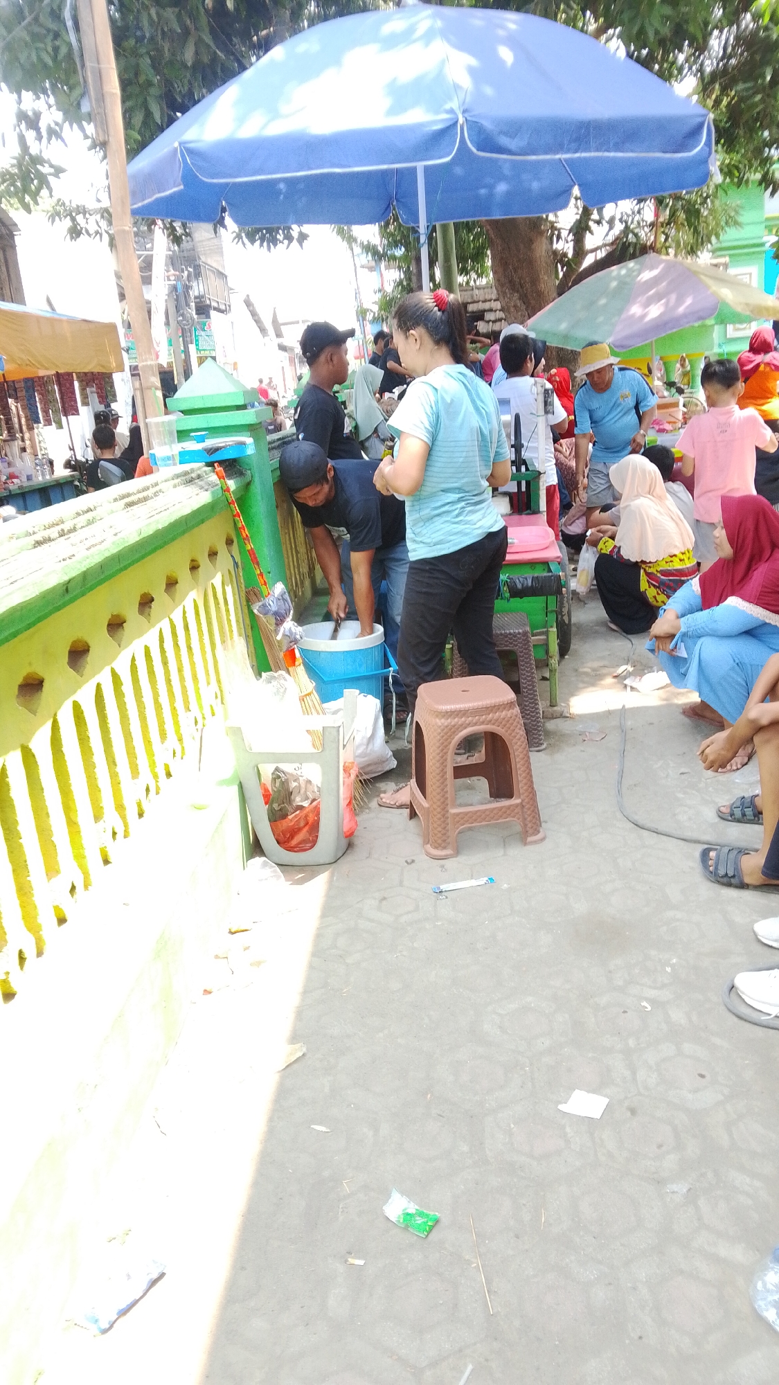 The image depicts a street scene in Mancar, Indonesia, under a large blue umbrella. Several individuals are present, engaged in various activities. In the foreground, a person in a dark t-shirt and black cap is bent over a blue plastic tub, appearing to be working with something inside it, possibly washing or preparing goods. To their right, a woman in a light blue t-shirt and dark pants stands, also looking towards the tub. A brown plastic stool is positioned between them.

Further back, a group of people are gathered near a cart or stall. Some are sitting on the ground, including a woman in a yellow and red patterned headscarf and a woman in a red headscarf and blue clothing. Others are standing, including a man in a blue shirt and a straw hat, and children in brightly colored clothing. The environment suggests a bustling outdoor market or a street-side food stall.

The ground is paved with hexagonal tiles, and various items are scattered on it, including trash and discarded wrappers. A green and yellow striped wall runs along the left side of the image. Trees with dense foliage provide shade, and parts of buildings are visible in the background. The overall impression is a lively and casual gathering in a public space.