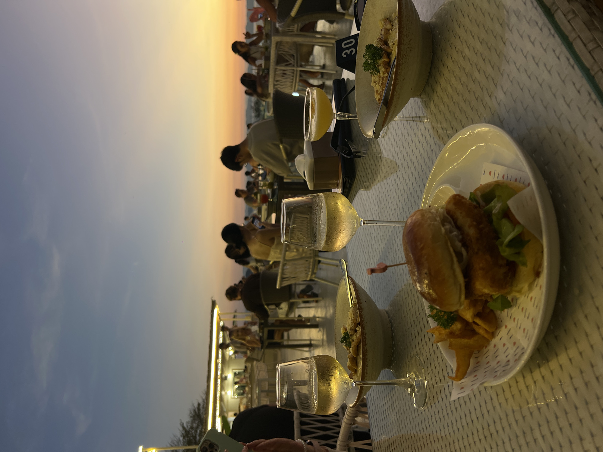 A nighttime image taken at a restaurant in Khao Thong, Thailand. The picture is focused on a table with a burger on a plate with fries and a bowl with food, with a partial number 30 visible. Two glasses of a white beverage sit near the edge of the table. Several silhouetted people are visible in the background with an orange-tinted sky in the distance. A structure is also visible in the lower-left of the frame.