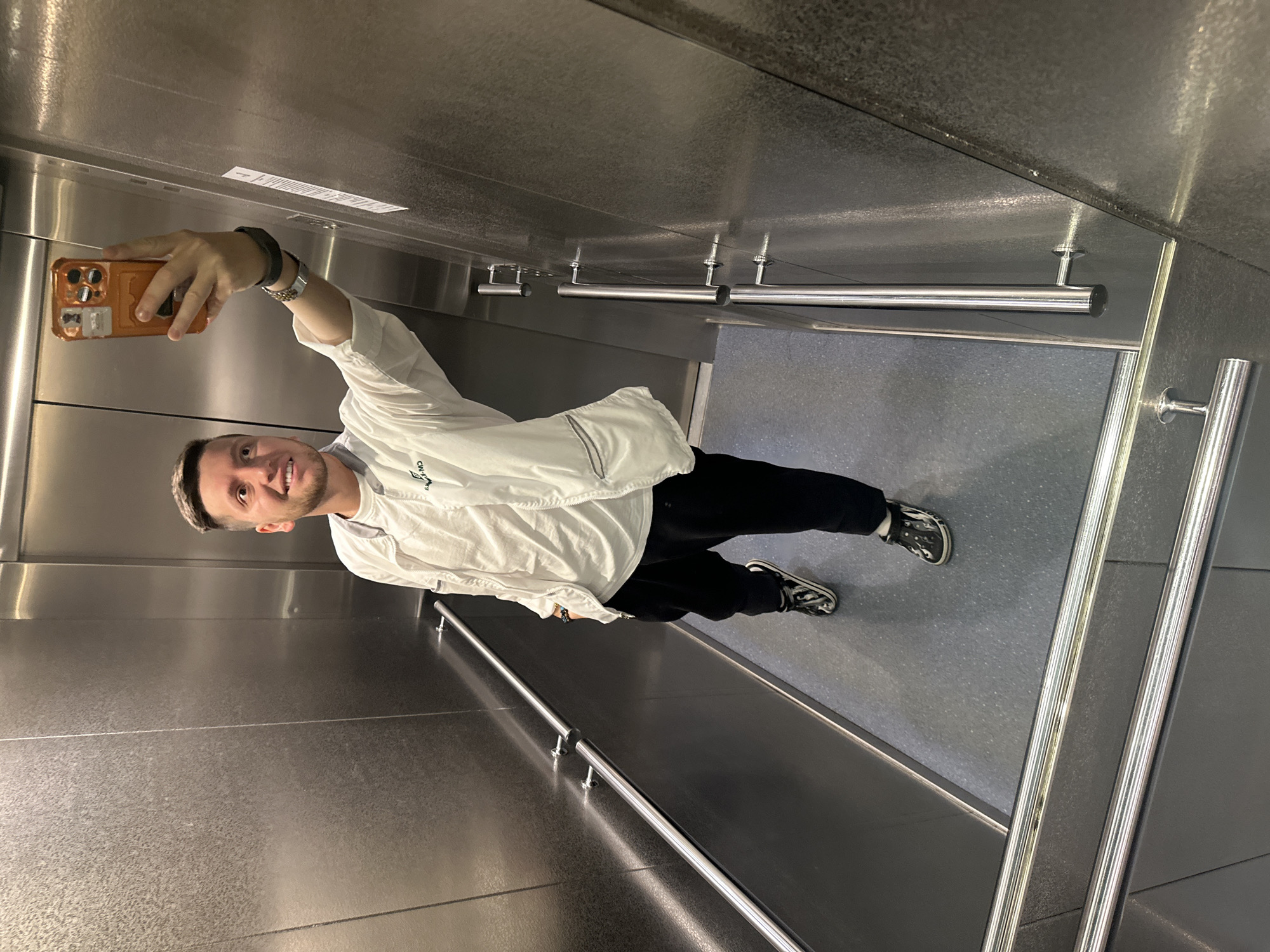 A man stands in an elevator taking a selfie. He is wearing a white jacket over a black shirt and black pants, with patterned black and white shoes. The elevator's interior is mostly metallic grey, with handrails visible. The elevator appears to be located in Moscow, Russia.