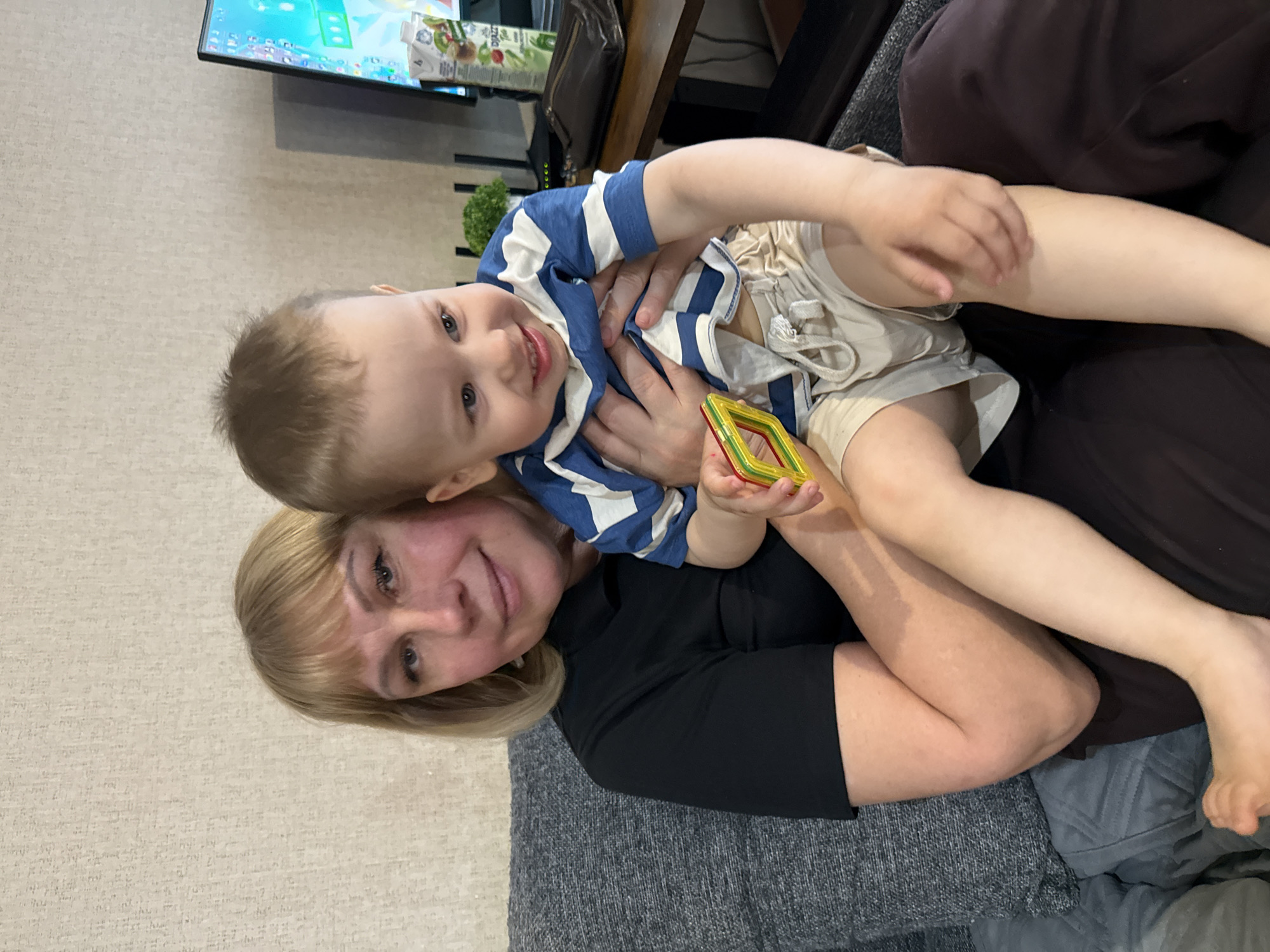 This heartwarming vertical photograph, taken in Rusanovo, Russia, captures an intimate moment between an adult woman and a young child. Both subjects are looking directly at the camera with genuine, joyful smiles.
The woman, with shoulder-length blonde hair and a warm expression, is seated on a dark gray upholstered sofa. She is wearing a black short-sleeved top and has her arms gently around the child, who is nestled closely against her.
The child, a toddler with light brown hair and bright eyes, is wearing a blue and white striped t-shirt and light-colored shorts. His smile reflects the woman's, conveying happiness and contentment. In his left hand, he holds a colorful magnetic building block, primarily yellow, red, and green, suggesting he was engaged in play.
The background reveals a home interior with a light-colored, textured wall. Above the subjects, a television screen displays vibrant, animated graphics, possibly from a children's program or game. Below the TV, a dark wooden shelf or cabinet holds a white juice carton. Visible text on the carton includes "FRUTTATO" and what appears to be "KIWI LIME MINT," along with illustrations of kiwi, lime, and mint leaves.
The overall scene radiates affection and a relaxed, casual atmosphere in a domestic setting. The lighting indicates it is likely daytime.