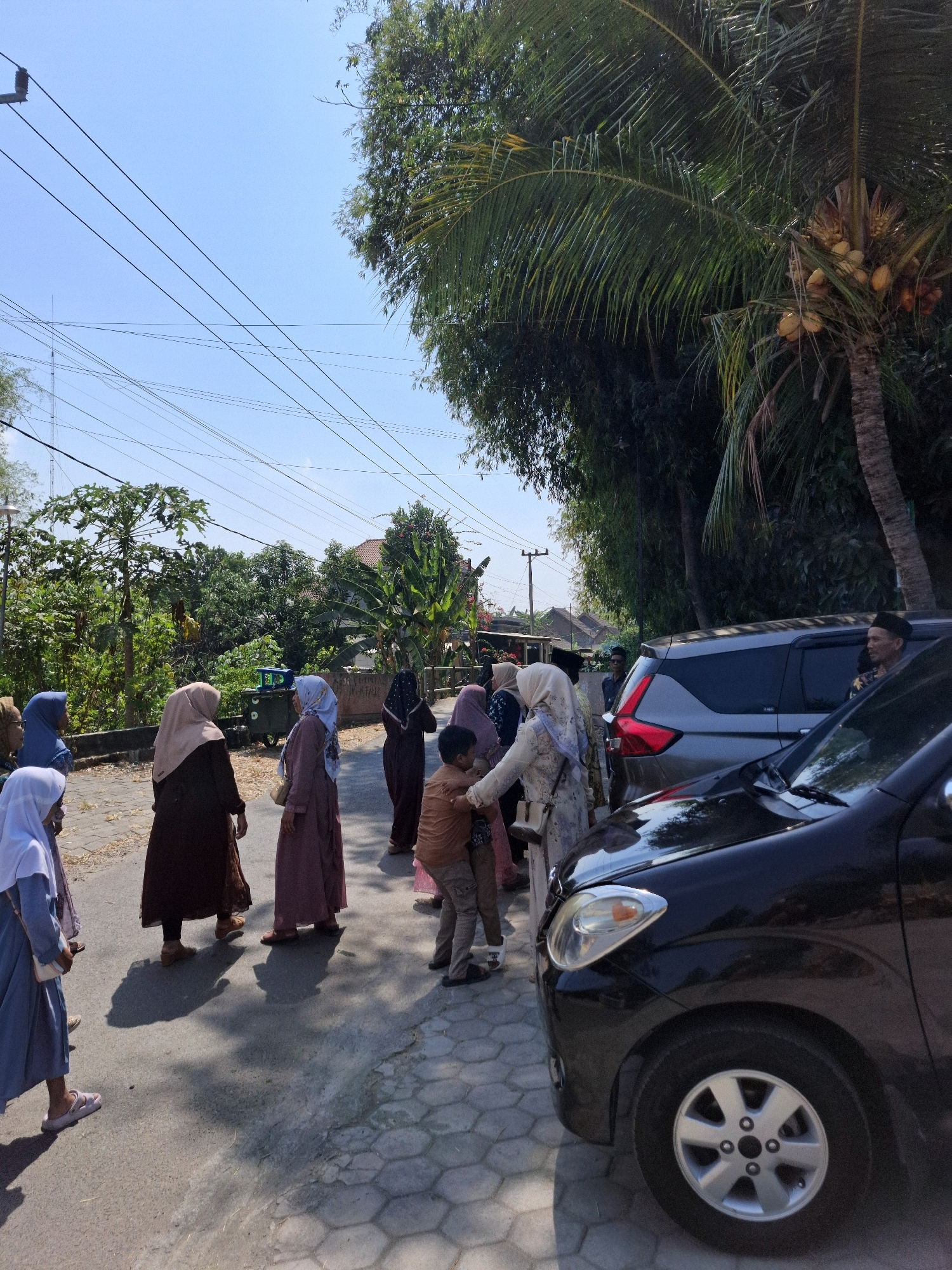 In the city of Kedungmaron, Indonesia, a group of people, mostly women wearing hijabs and modest clothing, stand on a paved street lined with trees and houses under a clear blue sky. A black car is parked in the foreground, and a silver SUV is parked behind it, partially obscuring a man in the driver's seat. Some individuals appear to be conversing, while others are observing their surroundings. The scene suggests a casual gathering or a moment of pause in a rural or suburban area. Coconut trees with visible coconuts are present in the background, adding to the tropical setting. Electrical wires crisscross the sky, indicating the presence of infrastructure.