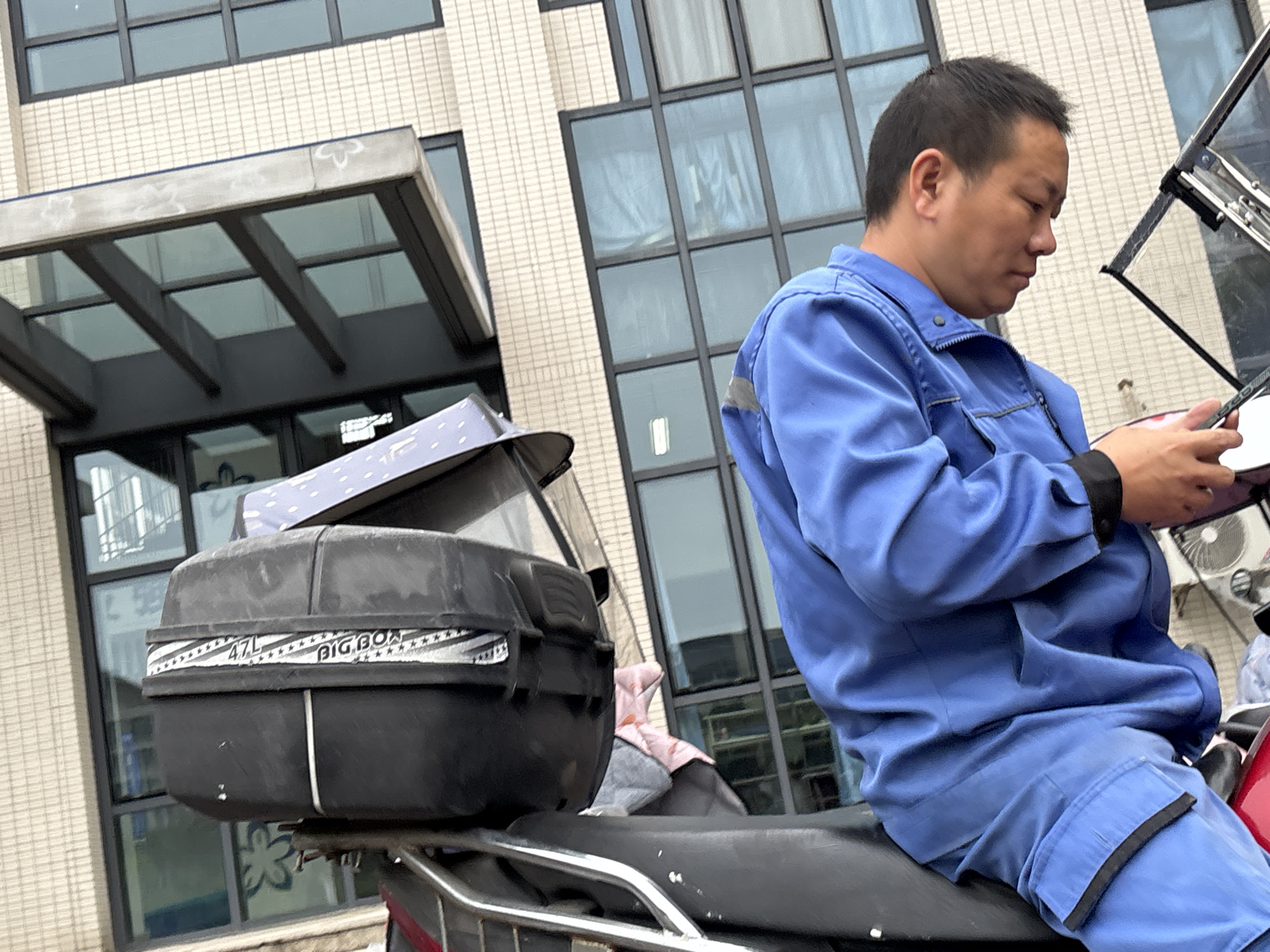 The image captures a man sitting on a motorcycle in Jiangjin, China. He is wearing a blue uniform and looking at a mobile phone in his hands. Behind him is a large black storage box attached to the motorcycle, labeled "BIG BOX 47L". The background includes a building with multiple windows and a glass awning. The scene is set during daylight hours.