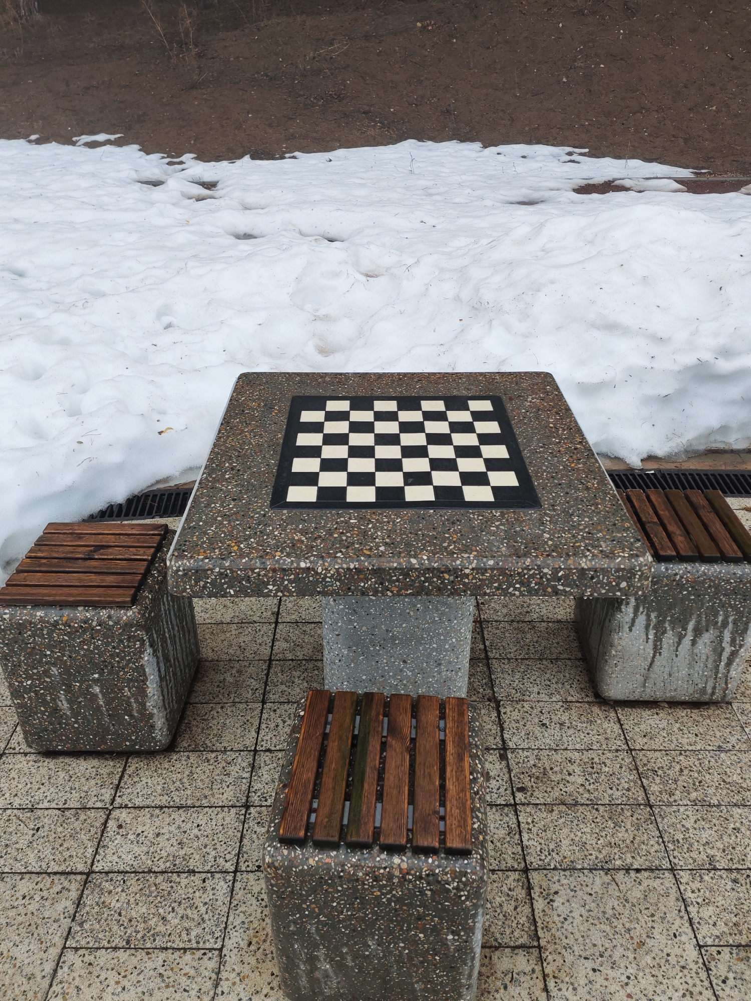 The image shows a concrete chess table with attached concrete benches in a partially snowy outdoor setting. The table has a square black and white chessboard inlaid on its surface. There are three benches: one on each side, with wooden slats as seating. The ground is paved with square concrete tiles. In the background, there is a slope covered with snow and dirt. The location is Chaikovsky, Russia. There are no people visible in the scene.