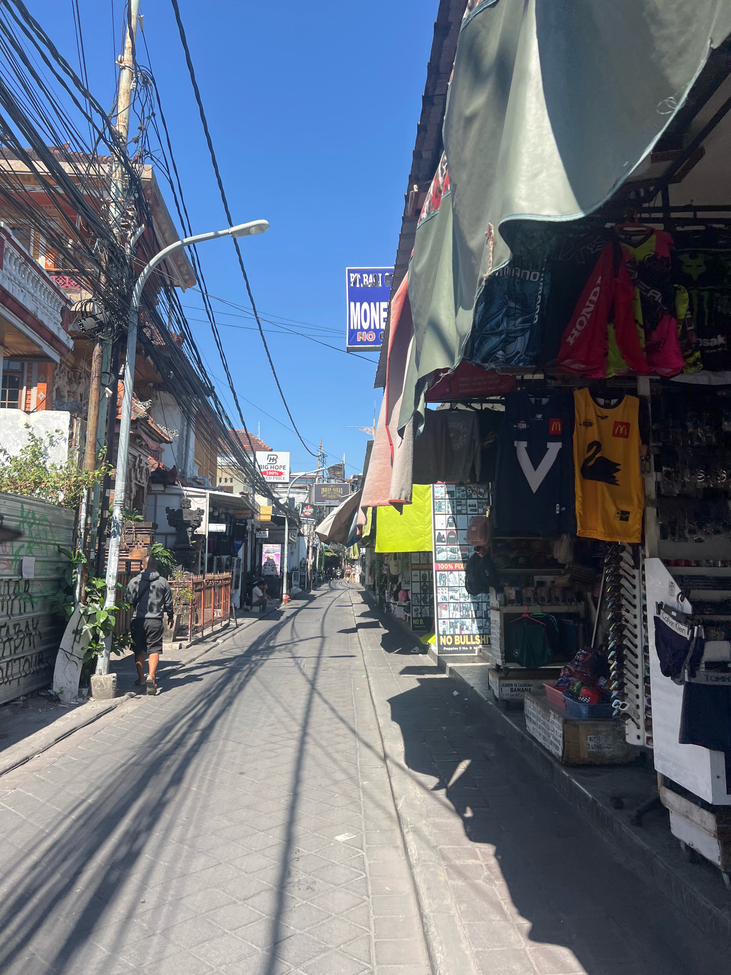 A sunny, narrow, cobblestone street in Kuta, Indonesia, is lined with shops displaying various merchandise. A person in a grey hoodie and black shorts walks away from the camera towards the background. Overhead, a dense network of tangled electrical wires crisscrosses the sky. Shops on the right sell clothing, including t-shirts and what appear to be sports jerseys, one yellow with a black swan logo and another dark blue with a white 'V'. Other shops on the left display more clothing and various trinkets. Signs are visible, including one blue sign that reads "MONEY" and "NO G". A large plant with green leaves grows to the left of the pathway. The scene depicts a bustling commercial street under a clear blue sky.
