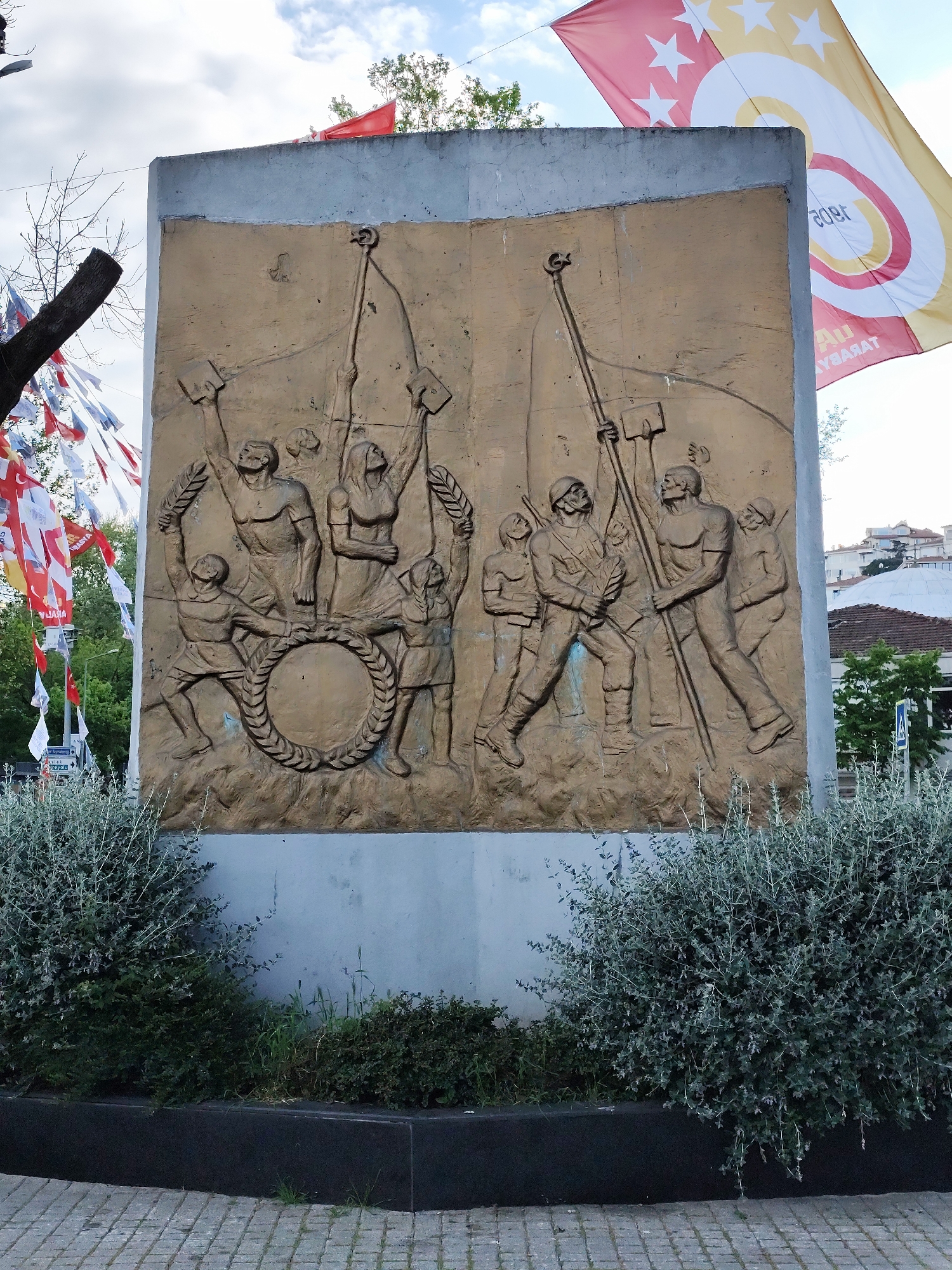 The image depicts a monument featuring a bas-relief sculpture in an urban setting, possibly a park or public square. The sculpture, set against a rectangular backdrop of concrete, is a brown-toned relief of human figures depicted in heroic or celebratory poses. On the left side, the figures are holding up a wreath, with some individuals holding aloft what appear to be laurel branches or leaves. On the right side, figures in military or worker-like attire are depicted holding what appear to be tools or implements and a flag. Above the monument, flags are visible, including one with what seems to be the Galatasaray sports club logo. The monument is surrounded by dense, low-lying bushes that border the base, which is made of a dark, polished material. The foreground shows a section of paved ground, indicating a walkway or plaza. The background consists of partial views of buildings, trees, and various flags hanging in the periphery, which are likely related to local events or celebrations. The location is Unknown, Unknown.