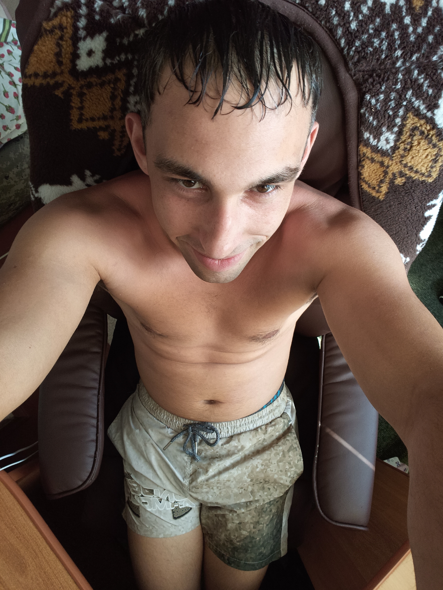 A man is lying down on a chair, with a blanket with a patterned design behind him. He is looking up and smiling at the camera, with his hair wet and slicked back. The man is shirtless, and wearing light-colored shorts with a camouflage pattern. The scene appears to be indoors, in a room with a wooden table in the foreground. The lighting suggests it is daytime.