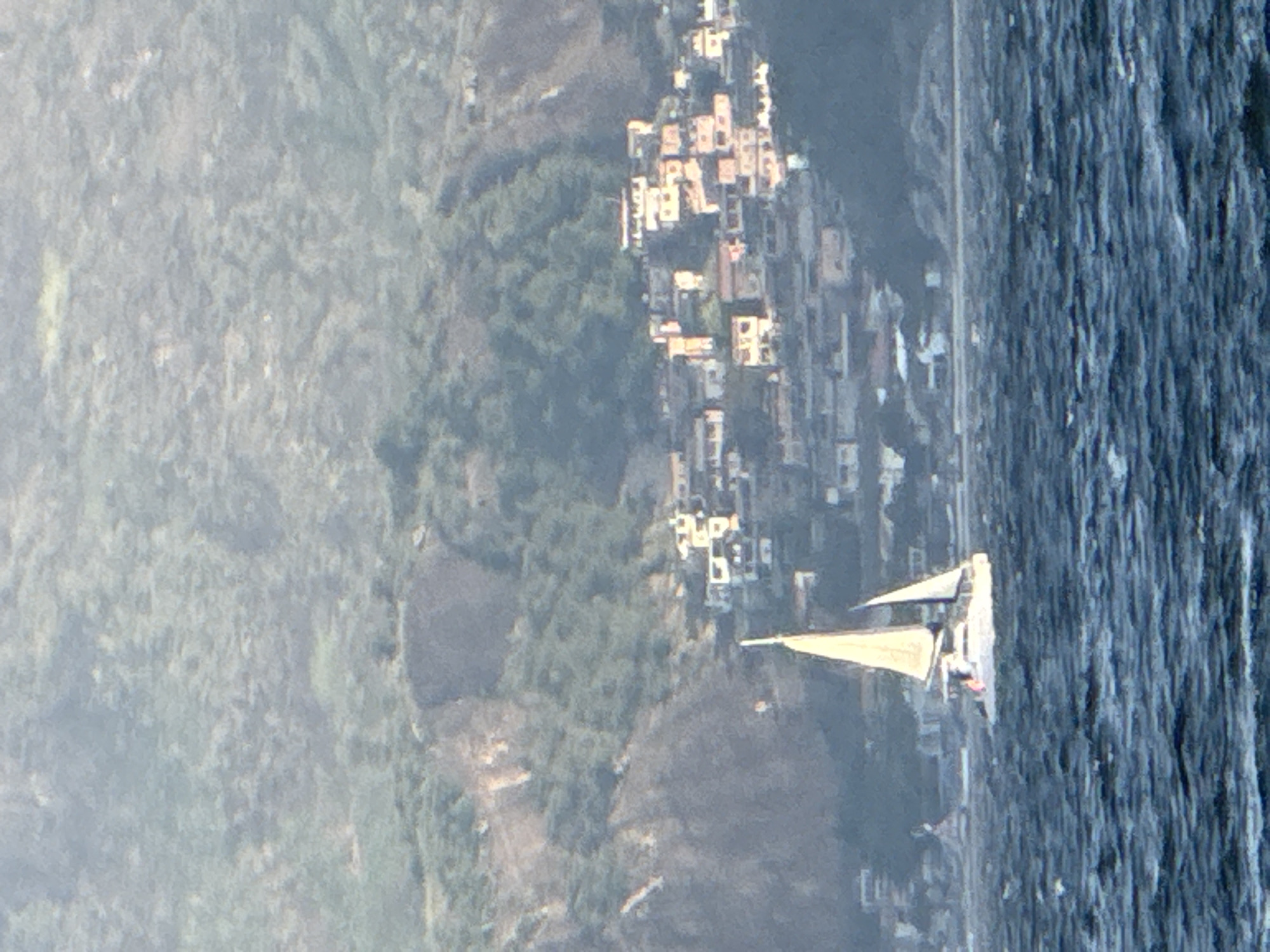 A catamaran sails on choppy, dark blue water with a densely packed hillside community visible in the background. The community is situated on a steep, green, and rocky incline. Buildings are closely clustered together, with many featuring light-colored walls and red or orange tiled roofs. The catamaran, a white vessel with two hulls, has its main sail and a smaller jib sail deployed. The sails are a light beige or yellowish color. The water is agitated, with visible wave crests and troughs. The overall lighting suggests it may be late afternoon or early morning, with a soft, diffused light. The scene appears to be the Guanabara Bay, with the hillside community likely being a favela or a densely populated residential area in Niterói, Brazil, which is known for its steep terrain overlooking the bay.