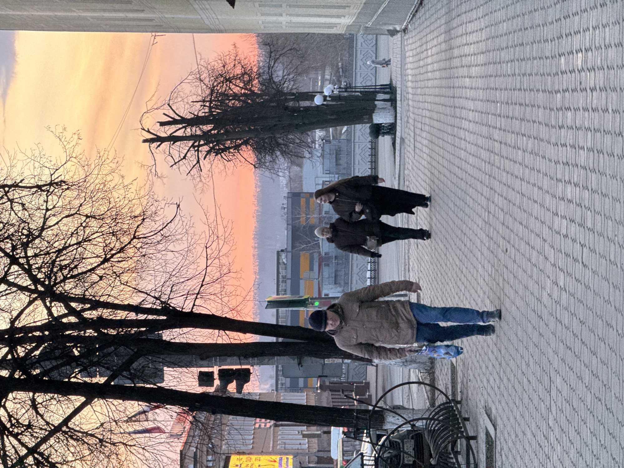The image depicts a street scene in Votkinsk, Russia, at either sunrise or sunset, suggested by the warm, orange-pink sky visible above the urban landscape. Several leafless trees with dark branches frame the left side of the scene. The street is paved with light gray bricks.
People are walking along the sidewalk. A man in a brown jacket, blue jeans, and a dark beanie is walking towards the camera. Two women are walking further up the street, away from the camera, wearing dark coats. In the distance, a lamppost and a figure are visible.
Buildings are visible in the background. A sign with Asian-looking lettering is seen through the trees. Cars are also parked on the side of the street.