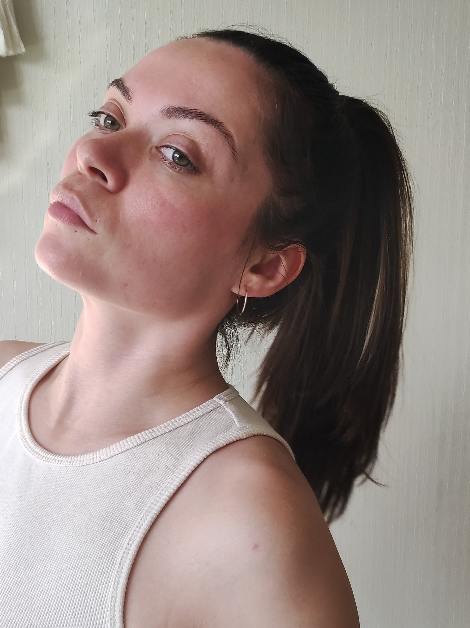 A light-skinned woman with dark hair pulled back in a ponytail poses for a photograph. She is wearing a light-colored tank top and a small gold hoop earring. Her gaze is directed upward and to the left. The background appears to be a plain, light-colored wall. There is no evidence to suggest that the location is Chaikovsky, Russia, or any specific city.