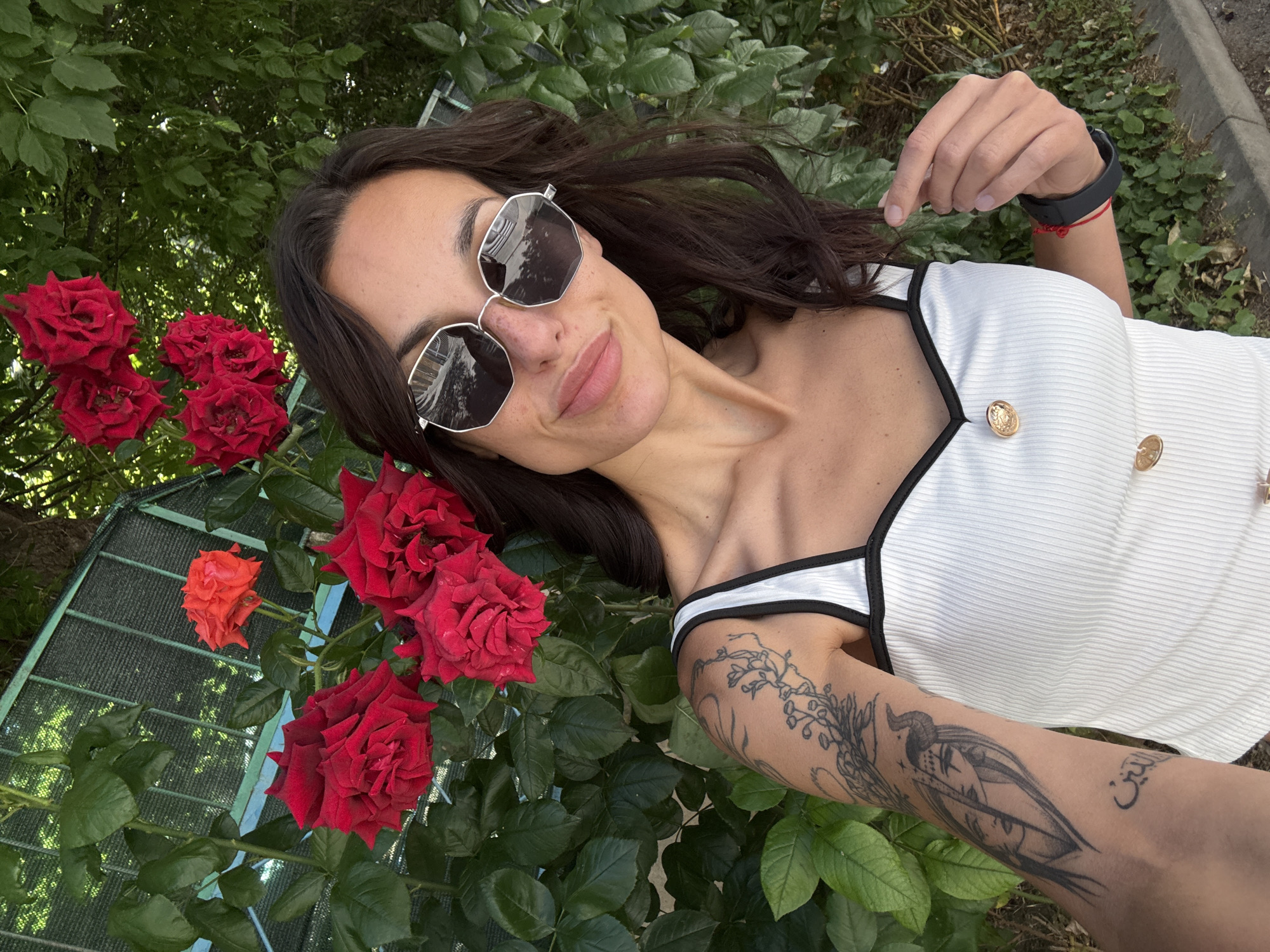 A woman with dark hair, wearing sunglasses and a white top with black trim, takes a selfie in front of a rose bush in Pleven, Bulgaria. She has tattoos on her left arm and a red string bracelet on her right wrist. The roses are red and orange, and the background shows green foliage and a fence.