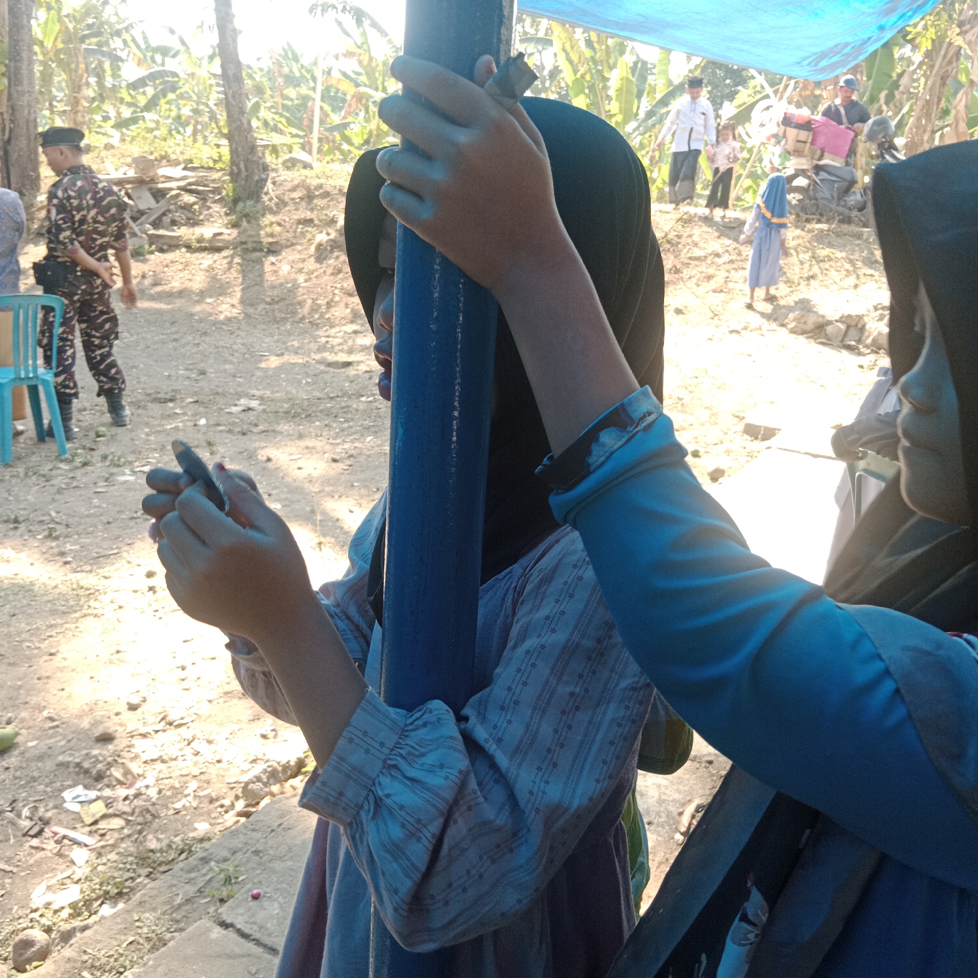 The image depicts an outdoor scene in Tegowanu Kulon, Indonesia. In the foreground, two individuals are partially visible, both wearing hijabs. One person, on the left, is wearing a blue shirt with a patterned fabric and a dark hijab. Their left hand is near their face, holding a small, dark object, possibly a pen or stylus. The other person, on the right, is wearing a bright blue garment and a black hijab. Their arm is extended, and their hand is also visible. Between them, a blue pole is prominent.
In the background, a person in a camouflage uniform, likely a soldier or security personnel, is walking to the left. They are wearing a black beret and a belt with pouches. Further back, several other people are visible, including a person in white clothing and a child in a pink and white outfit, standing near a motorcycle loaded with items. Another child in a blue hijab is walking away from the camera. The environment is rural, with trees, vegetation, and a dirt or gravel ground. A bright blue tarp is stretched overhead, providing shade. The lighting suggests it is daytime, with sunlight casting shadows. The overall atmosphere appears to be a gathering or event in a public outdoor space.