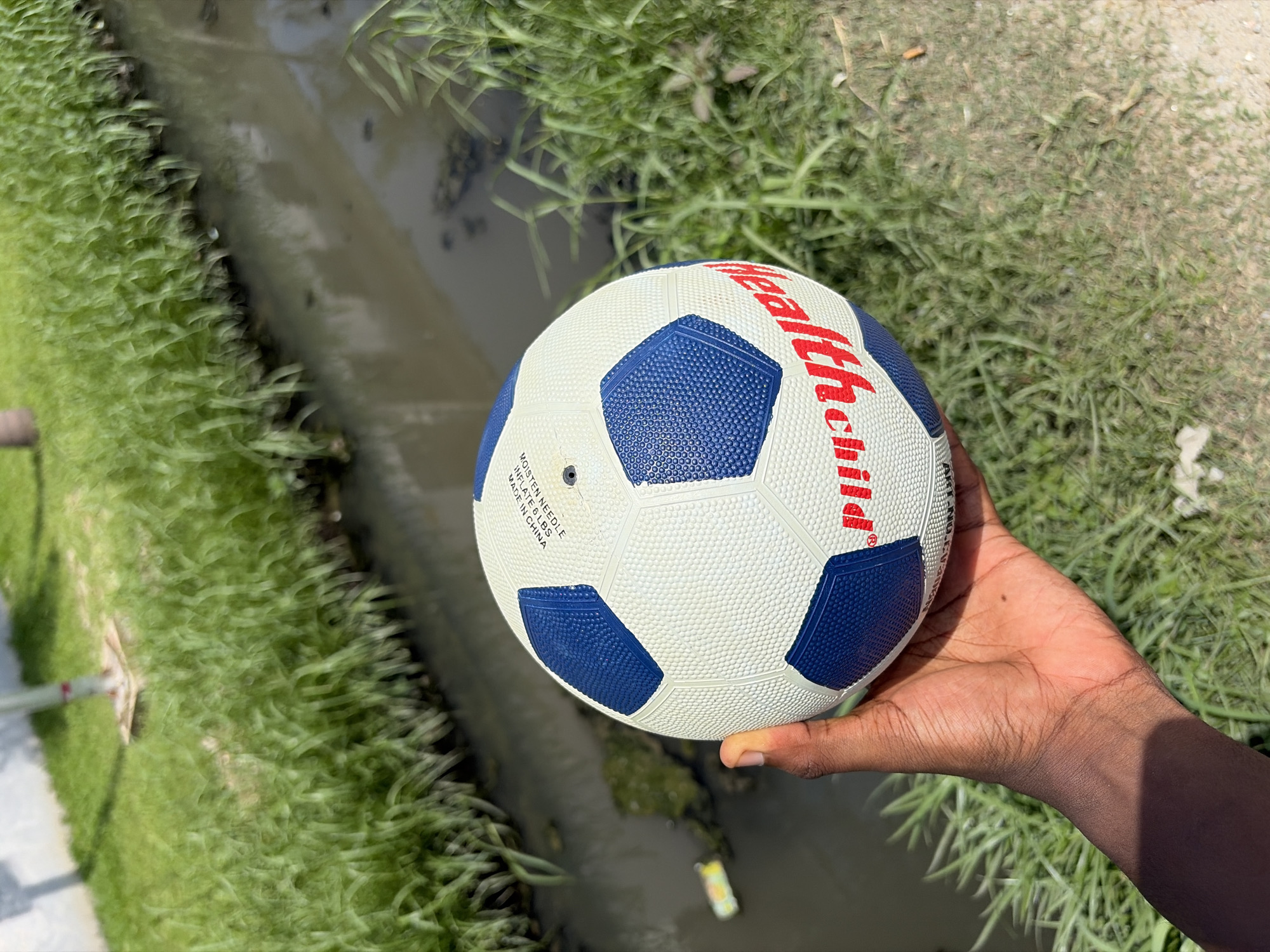 The image shows a person holding a blue and white soccer ball with red lettering that reads "healthchild" in their right hand. The ball is positioned in front of a grassy area adjacent to a narrow stream or ditch filled with water. The location appears to be outdoors, with vegetation surrounding the water source. No specific details identify the location as Alaguntan, Nigeria.