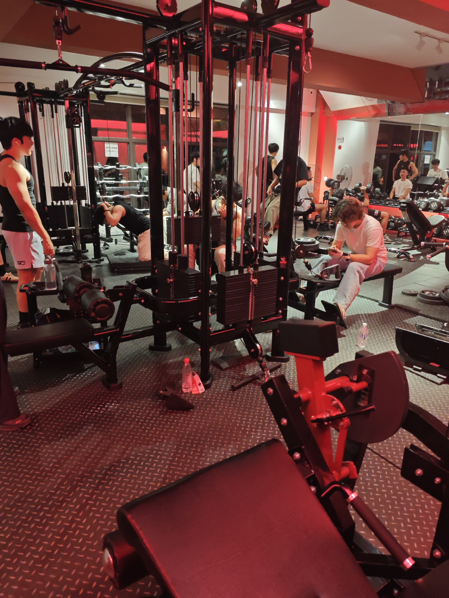 The image depicts the interior of a gym located in the Chongqing High-tech Zone, China. Multiple individuals are present, engaged in various activities. One man, wearing a black tank top and light shorts, stands holding a water bottle. Another man in a black tank top kneels, seemingly stretching. In the background, several other individuals are working out with weights and machines. One person is sitting on a bench while looking at their phone while wearing a white t-shirt. The gym features various weight training machines, including cable machines and leg press equipment. The floor has a textured metal surface, and the lighting is primarily red. Water bottles are visible on the floor near some of the equipment.
