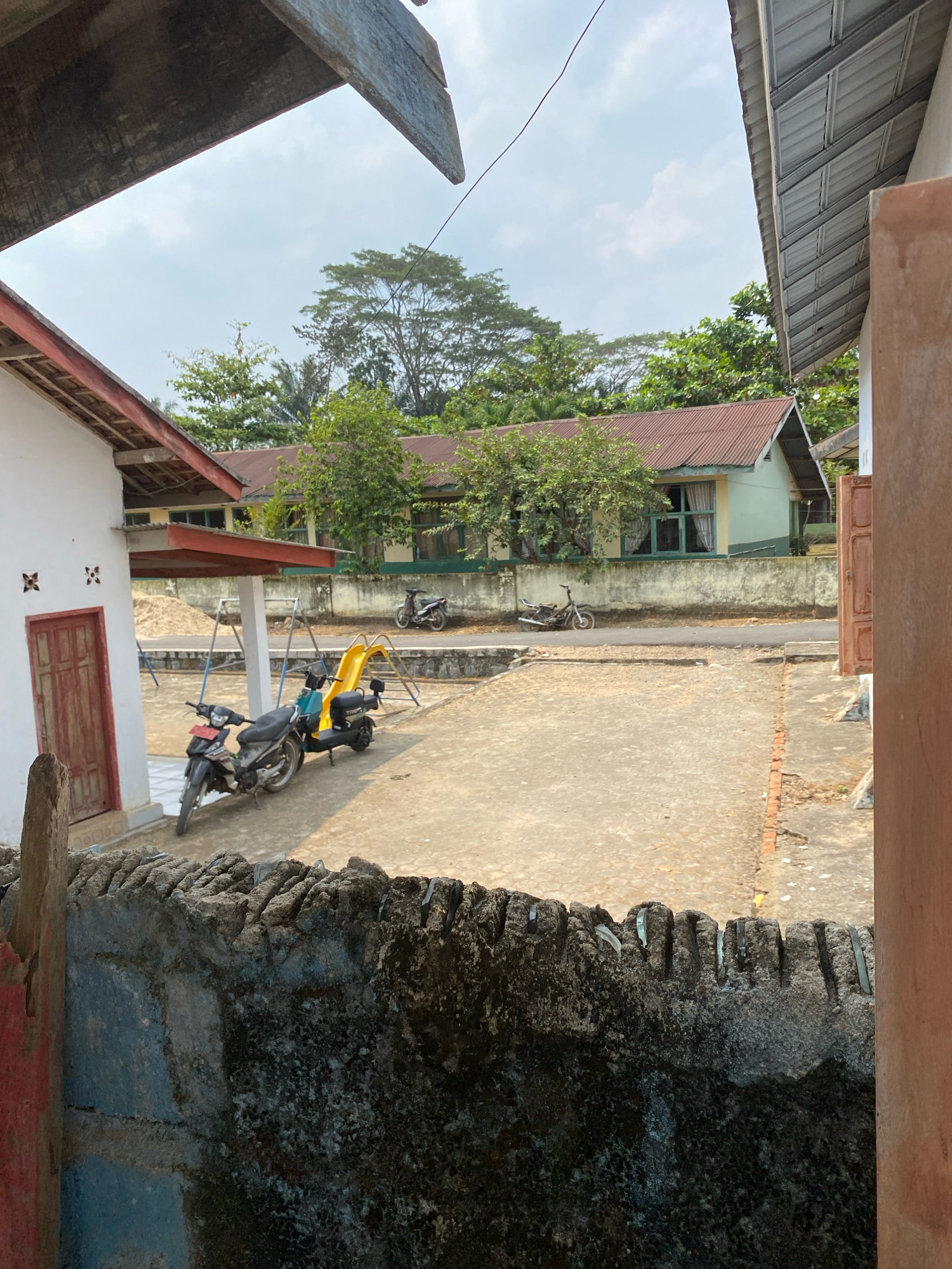 The image depicts an outdoor scene, likely in a residential or institutional area in Mojo Pahit, Indonesia. In the foreground, a rough, grey, and moss-covered wall with broken glass shards embedded along its top edge runs horizontally. To the left, a portion of a red wooden post and a section of a white building with a red-framed door are visible.
The midground reveals a paved courtyard area. Two motorcycles are parked near a bright yellow slide with blue supporting poles, suggesting a children's play area. The motorcycles appear to be of a common commuter type, one dark-colored with a red license plate, and another black one. Further back, a light green building with a weathered brown roof and a row of windows with curtains is present. A concrete wall with a rough texture separates the courtyard from the building. Two more motorcycles are parked along the road in front of this building.
The background is filled with lush green trees and vegetation, indicating a tropical or subtropical climate. The sky is overcast with scattered clouds. The overall impression is one of a functional, somewhat aged, and lived-in environment.
