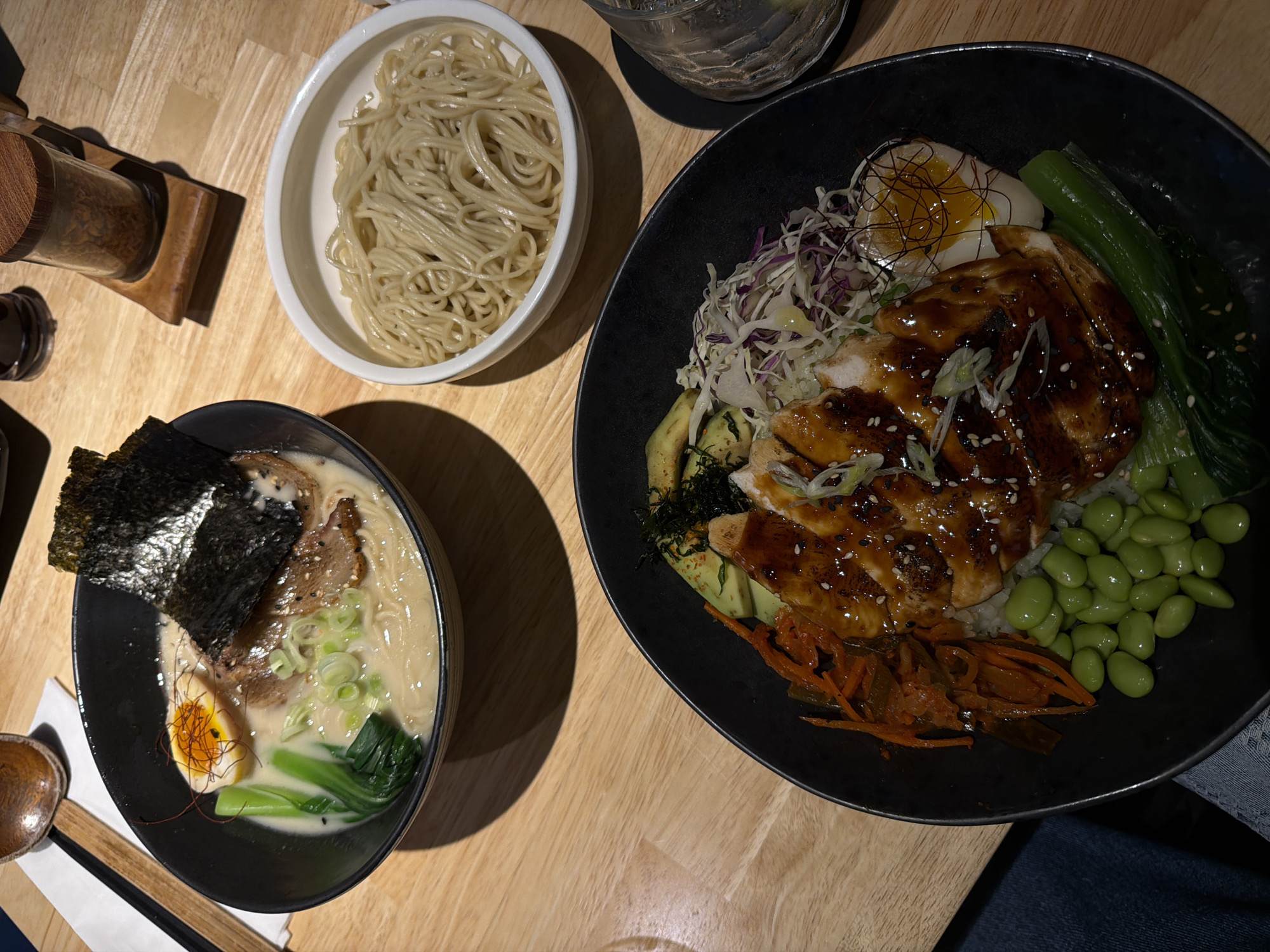 The image shows a wooden table with three dishes of Japanese cuisine. In the lower left is a bowl of ramen with a dark broth, topped with seaweed, meat, green onions, and a soft-boiled egg. Beside it are chopsticks and a spoon. In the top left is a container with a spice. In the upper middle is a white bowl filled with noodles. To the right is a black bowl containing rice, chicken, edamame, carrots, bok choy, avocado, purple and white cabbage, and a soft-boiled egg. Based on the style of food, it is likely that this image was taken in North Kuta, Indonesia.