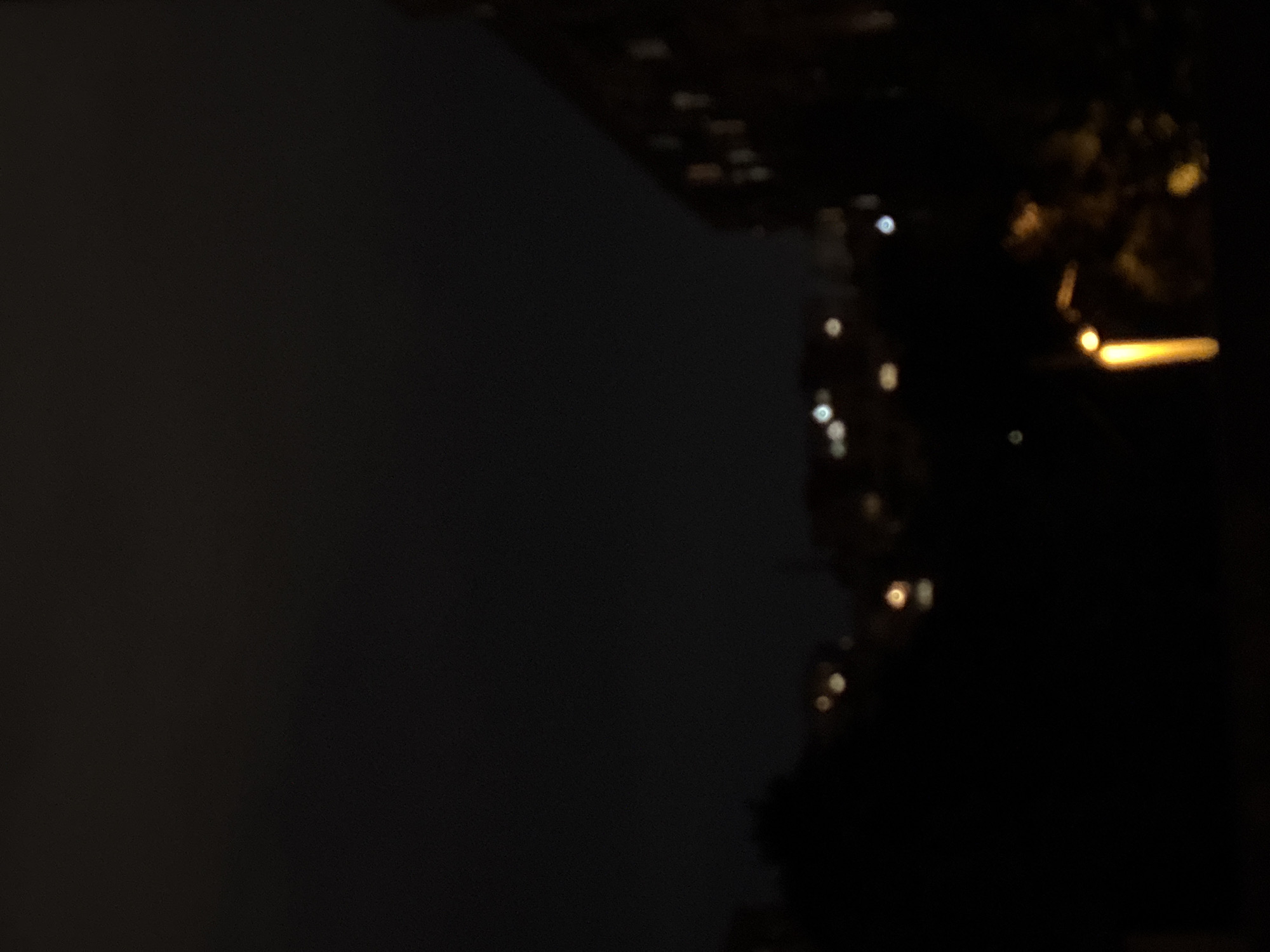 The image is a dark, out-of-focus photograph taken at night in an urban environment, likely a residential area in Sofia, Bulgaria. The scene is dominated by the blackness of the night sky. In the mid-ground, blurred shapes suggest the silhouettes of buildings and trees. Distant, out-of-focus lights, likely from windows or streetlights, create soft, circular bokeh effects. On the right side of the frame, a building's facade is vaguely visible, with faint rectangular shapes suggesting illuminated windows. In the foreground, a bright, vertical yellow light source, possibly a lamp or a streetlight, is prominent. The overall impression is one of a city at rest, shrouded in darkness, with only scattered artificial lights piercing the gloom.