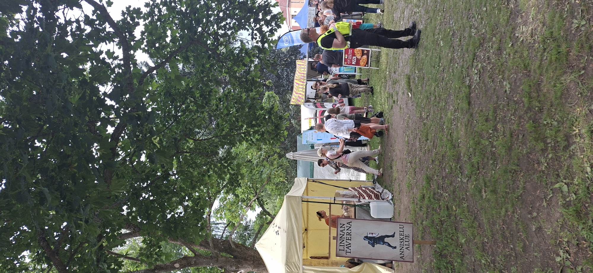The image depicts an outdoor event likely in Turku, Finland, with multiple stalls and attendees gathered under a large, leafy tree. In the foreground, a sign for "LINNAN TAVERNA" with an image of a knight is visible. To the right, a man wearing a bright yellow vest stands facing the right edge of the frame. Several stalls are set up, including one with a beige tent selling drinks and another further back with food items advertised. People are seen walking, browsing, and interacting. The ground is covered in grass and dirt. The overall atmosphere suggests a public gathering or festival.