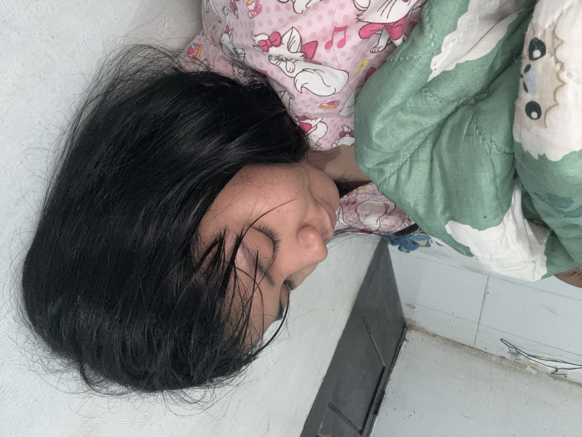 In North Jakarta, Indonesia, a young girl with black hair is sleeping on a floor covered with white tiles. She is lying on her side, partially covered by a green blanket with a cartoon sheep pattern and a pink pajama top adorned with cartoon cats and musical notes. The environment appears to be an indoor space with a white wall and a dark door frame in the background. There are also some wires and a clothes hanger visible on the floor. The girl's face is relaxed, indicating deep sleep.