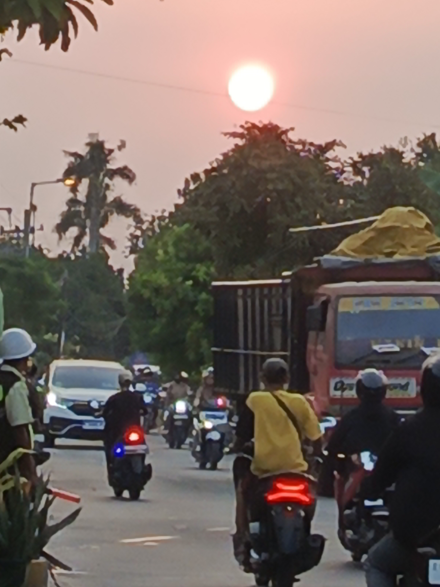 In the city of Wedi, Indonesia, a street scene unfolds at dusk. The sky, a soft pink, is dominated by the setting sun, a bright white orb casting a warm glow. A line of motorcycles, some carrying multiple passengers, navigates the road. One motorcycle in the foreground displays a red and blue taillight, possibly indicating law enforcement or emergency services. Behind them, a white car and a red truck with a yellow tarp-covered load are visible. Trees and streetlights line the roadside, framing the vehicular activity.