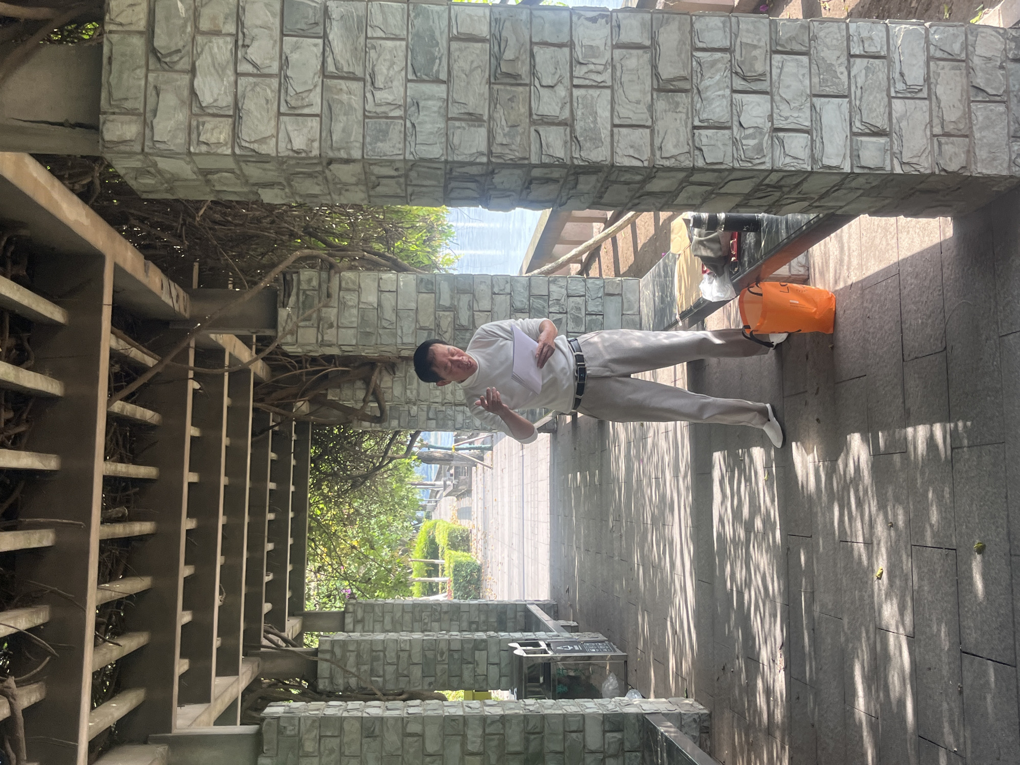 The image, correctly oriented, shows a man standing on a paved outdoor pathway under a stone and trellis structure in Jiangjin, China, on a bright, sunny day.
The central subject is an East Asian man, standing center-right, wearing a light-colored (likely white or light grey) long-sleeved polo shirt, light-colored trousers, and white shoes, with a black belt. He holds a stack of white papers or a thin book in his left hand, while his right hand is slightly raised, as if gesturing or speaking. His expression is focused.
He stands on a pathway paved with light grey rectangular tiles, which shows strong, dappled shadows from the overhead structure and foliage, indicating bright sunlight. To his right, an orange tote bag rests on the ground. Further back and to his left, beneath the overhead structure, is a metal trash can with a black lid. Visible text on the trash can includes Chinese characters and the English word "RECYCLE".
The man is positioned within a sheltered walkway or pergola, characterized by sturdy pillars and beams made of grey, rough-hewn stone blocks, and an overhead trellis-like structure made of metal or wood slats through which dense, tangled vines and branches grow. Beyond the immediate structure, lush green trees and foliage are visible. In the distant background, a body of water, likely a river or lake, can be seen under a bright sky. The pathway extends into the distance, flanked by landscaped greenery. The overall scene suggests a public park or a riverside promenade.