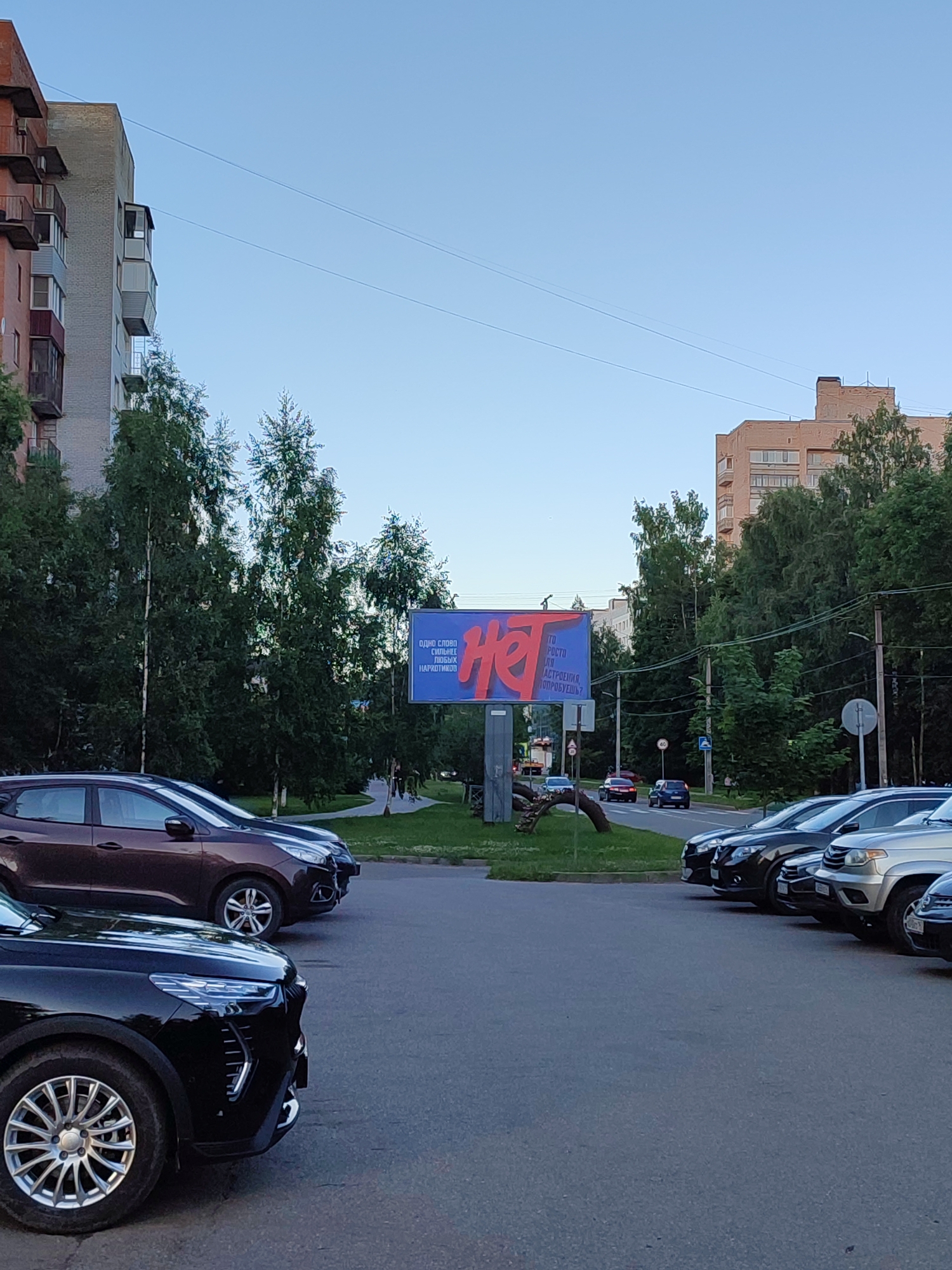 A billboard advertising "Нет" (No) is prominently displayed in Vsevolozhsk, Russia. The billboard is red and blue, with the word "Нет" in large red lettering. To the left of "Нет," smaller blue text reads: "Одно слово сильнее любых наркотиков" (One word is stronger than any drugs). The billboard is positioned in an urban area with apartment buildings and trees visible in the background. Several cars are parked in a lot in front of the billboard, and a street with moving traffic is in the foreground. The sky is clear and blue, indicating daytime.