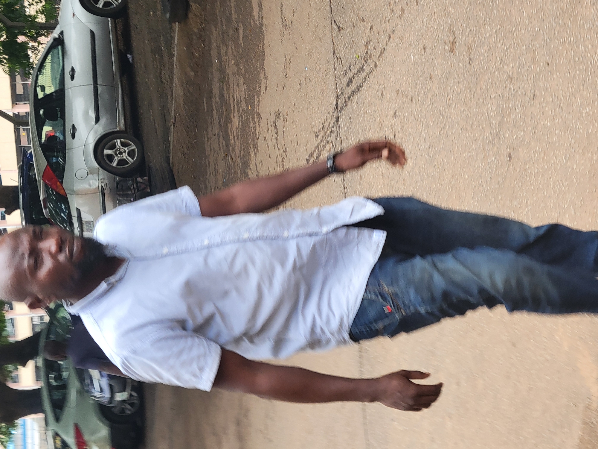A bald Black man with a beard walks toward the right of the frame, his body angled away from the camera. He wears a light blue, short-sleeved button-down shirt and blue jeans. The man's left arm is bent at the elbow, with his hand resting on his hip. A watch is visible on his left wrist. The background includes a silver hatchback car with a dented rear bumper and a tree casting shadows on the ground. The scene appears to be in an outdoor urban setting, possibly a parking lot or street in Abuja, Nigeria, given the presence of vehicles and a somewhat unkempt environment with cracked pavement.