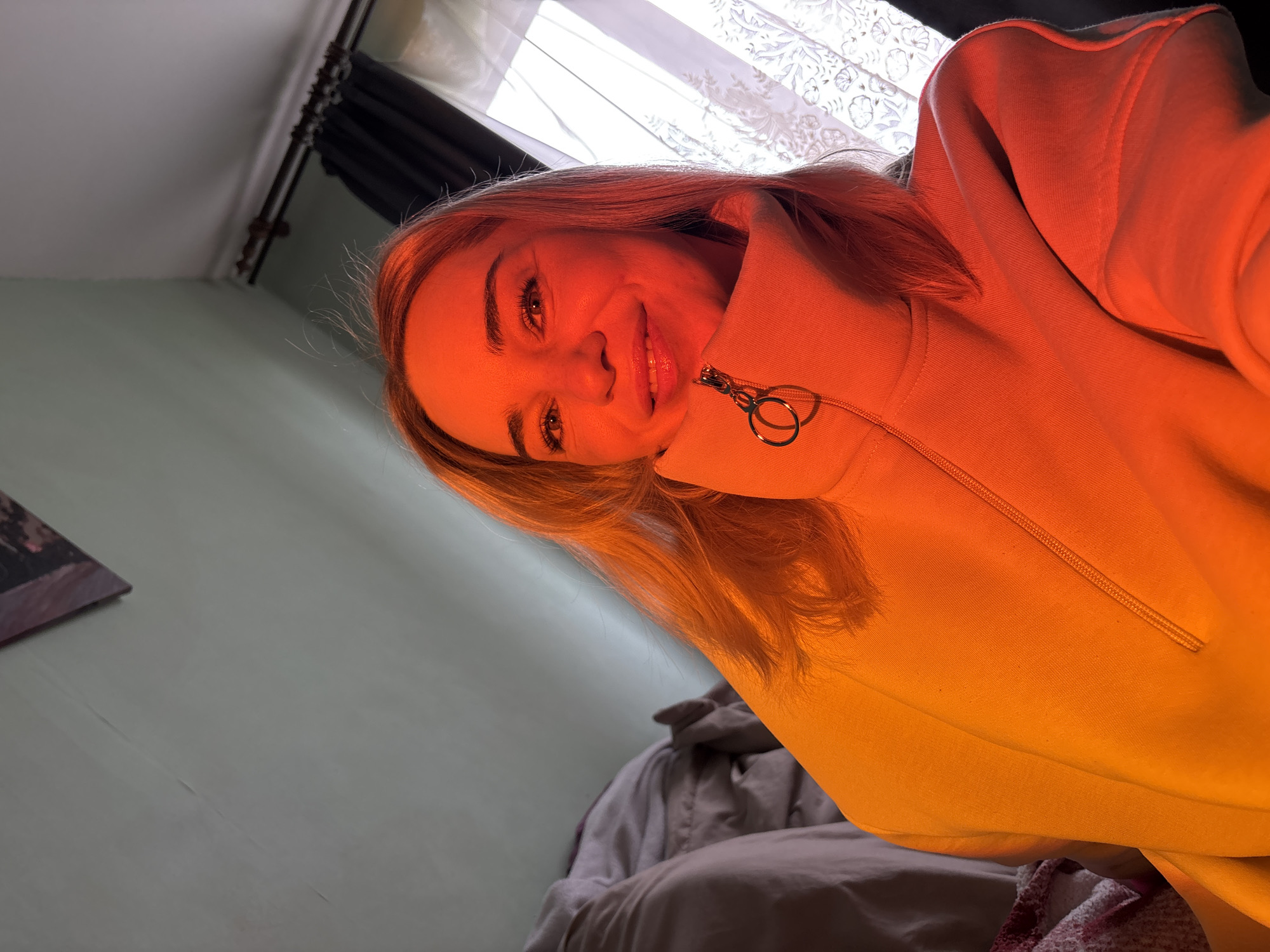 A selfie-style photo captures a woman with light blonde hair, possibly taken indoors. Her face is illuminated with a strong orange glow, which also affects the color of the zip-up hooded sweatshirt she is wearing. She is smiling slightly at the camera. The background suggests an interior setting, with indistinct shapes that might be furniture and parts of a window with curtains visible. A glimpse of a wall is visible behind her. The location is not apparent from the image details provided.