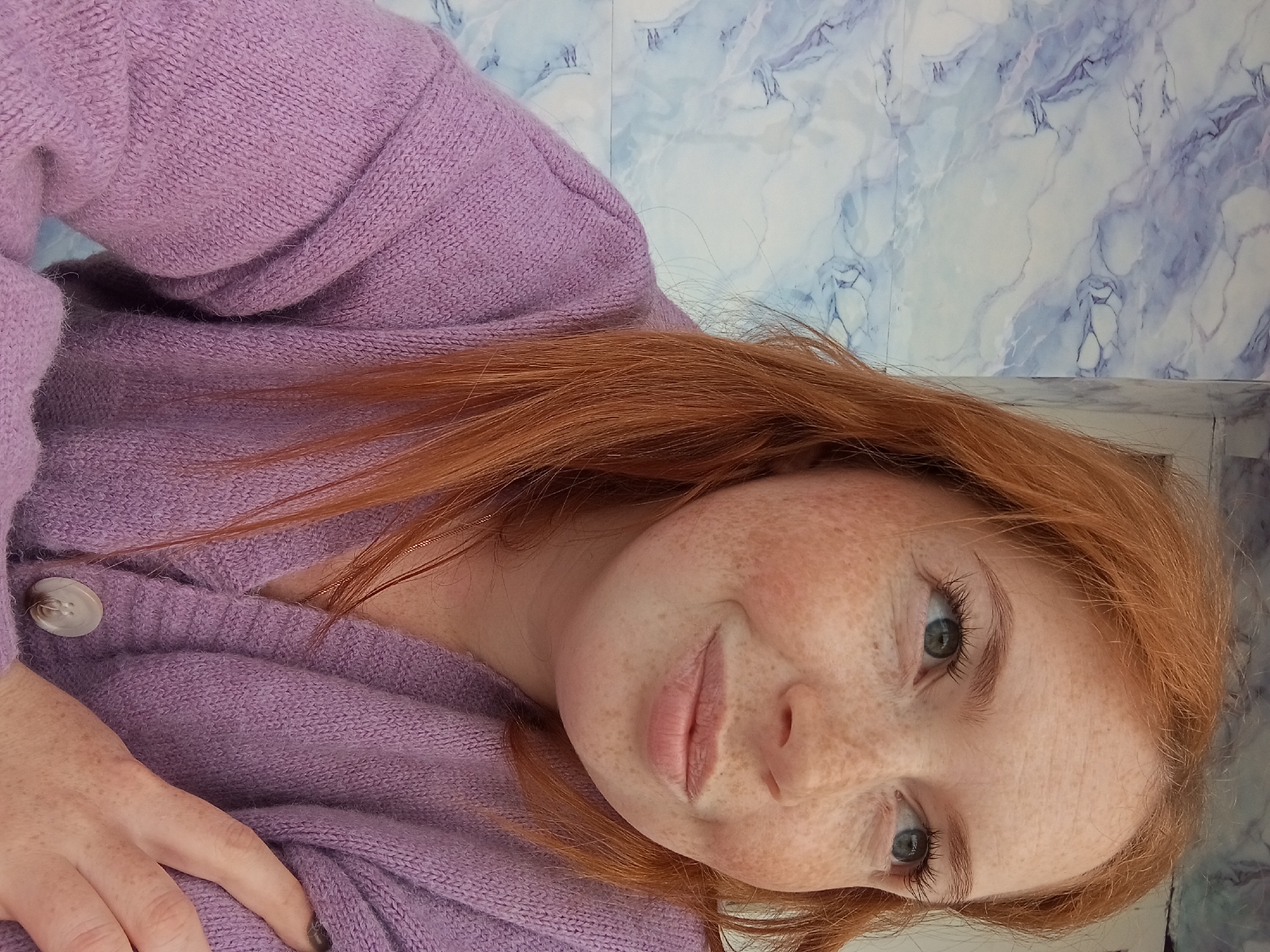 A young woman with red hair and numerous freckles on her face and visible hand is depicted in a close-up, chest-up portrait. She has light-colored eyes, appearing greenish-blue, and a subtle smile. Her red hair is somewhat dishevelled, framing her face and falling over her shoulders. She is wearing a soft, knitted or woven light purple (lavender) sweater or cardigan, which features a visible button on the front near her left hand. Her left hand, also freckled, rests gently on the sweater, and one fingernail appears to have dark polish.
The woman is looking directly towards the camera, her head slightly tilted. The lighting suggests daytime. The background behind her is a wall or surface with a distinct marbled or veined pattern, predominantly white and light blue, resembling natural stone or a decorative tile. The overall scene conveys a casual, personal moment. There is no visible text in the image. The location context provided is Yurgamysh, Russia.