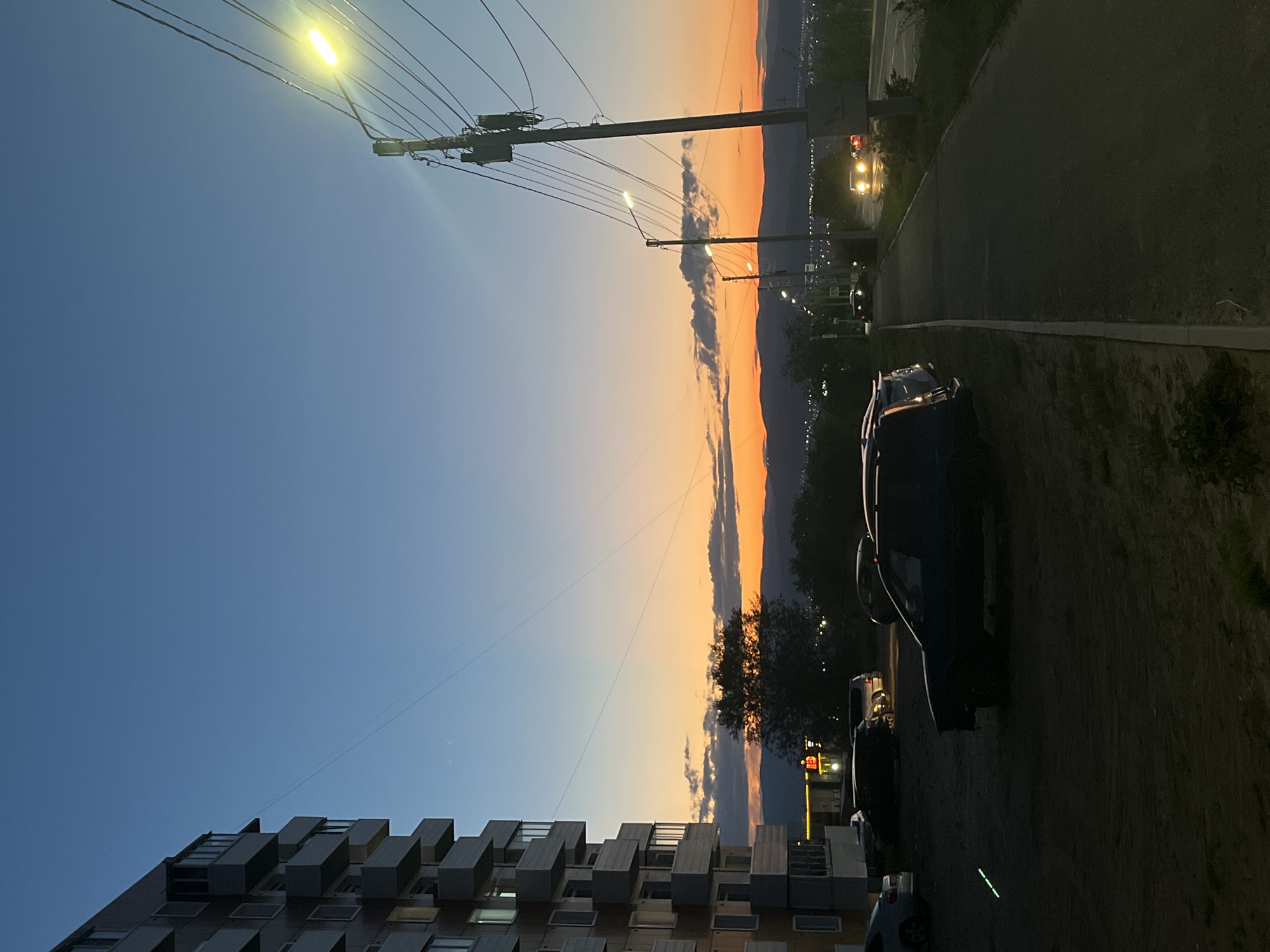 The image depicts an urban scene at dusk, characterized by a gradient sky transitioning from deep blue at the top to vibrant orange and yellow near the horizon. On the left, a multi-story apartment building with visible balconies dominates the foreground. To the right of the building, a street with a sidewalk is visible, lined with utility poles and streetlights. Several cars are parked in a lot in front of the building and along the street. In the distance, a hazy mountainous silhouette is present, with city lights faintly visible. The overall ambiance is one of twilight in a residential area.