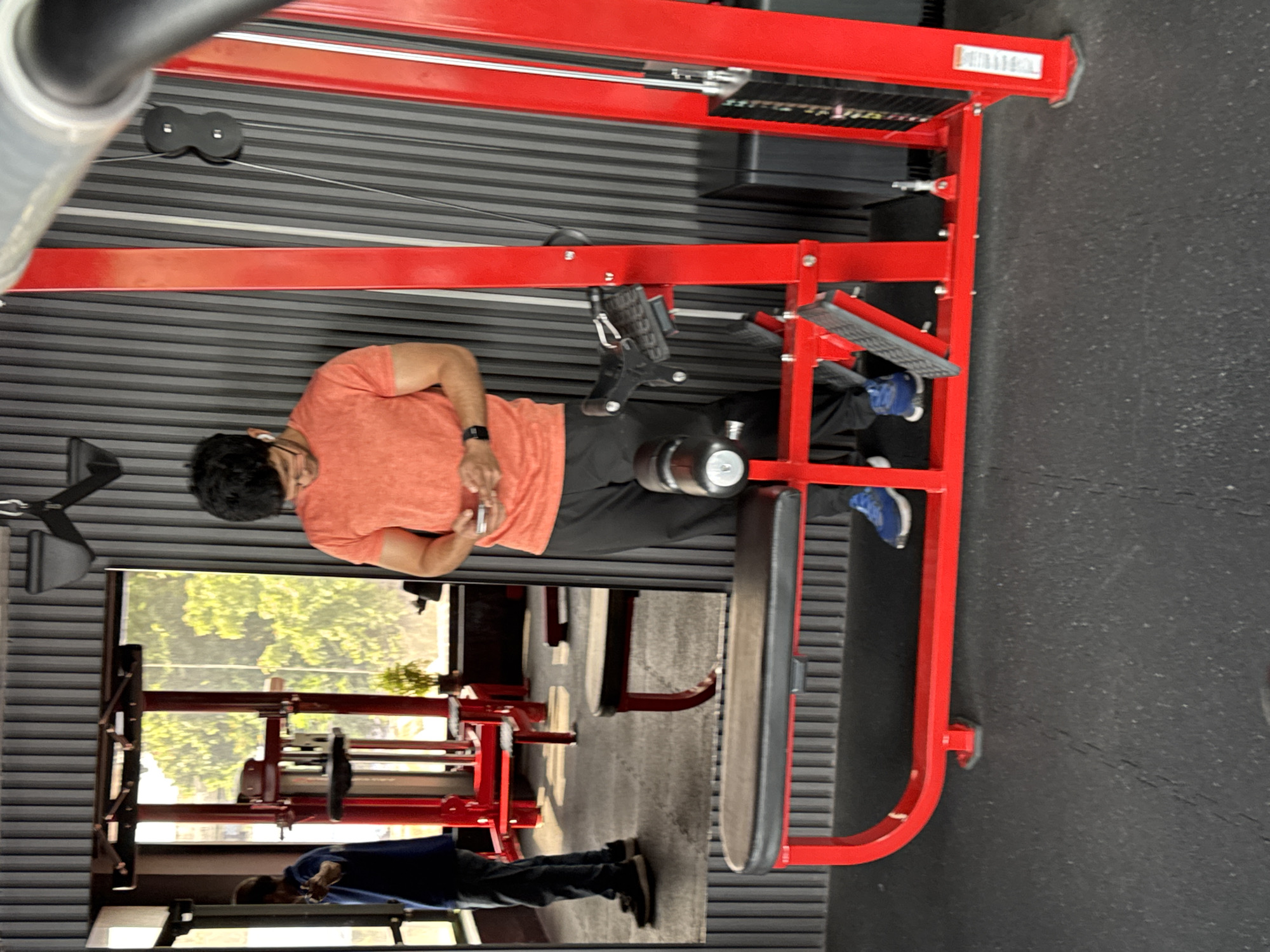 A man is standing in a gym in Kolkata, India, using his phone. He is wearing an orange t-shirt and black pants. He is standing next to a red weight machine. In the background, there is a mirror reflecting another person and more gym equipment. The walls are a dark ribbed texture. The floor is black rubber matting.