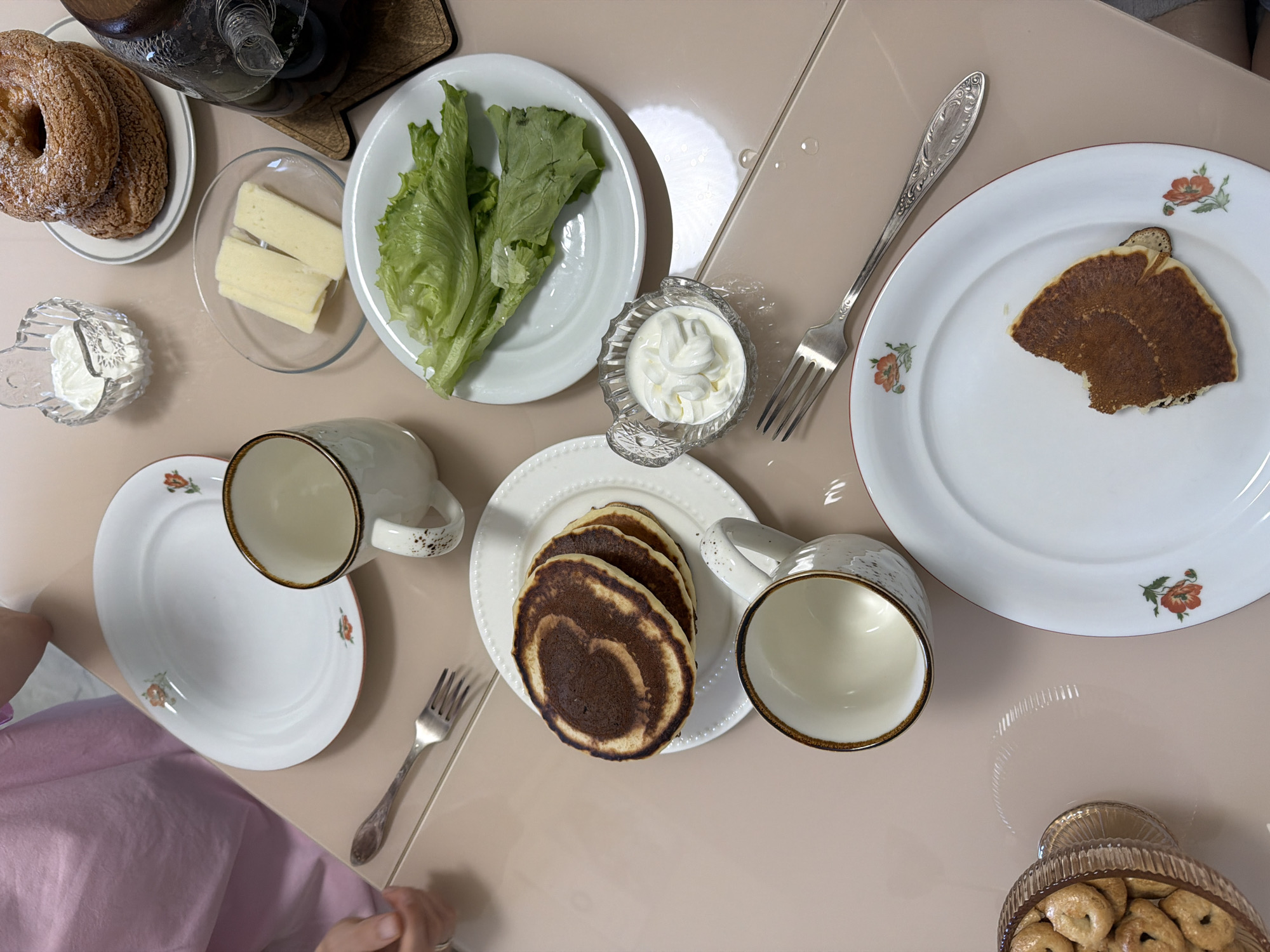 A top-down shot shows a table set for a meal, possibly breakfast or brunch. There are various food items arranged on the table: pastries, pancakes, cheese, salad, and small bagel-like snacks in a footed glass dish. The pancakes are round with a darker center, and some are stacked on a white plate. There's a partially eaten pancake on another plate. Two white mugs are placed near the plates with pancakes. Cream is in a small, ornate glass dish. Silverware is present, consisting of forks placed near plates. A teapot and another dish are at the top left of the frame. A person wearing a pink garment is partially visible on the left side of the image. The table surface is smooth and light brown. The scene appears indoors in a residential setting. There is no information to determine the location as Izhevsk, Russia.