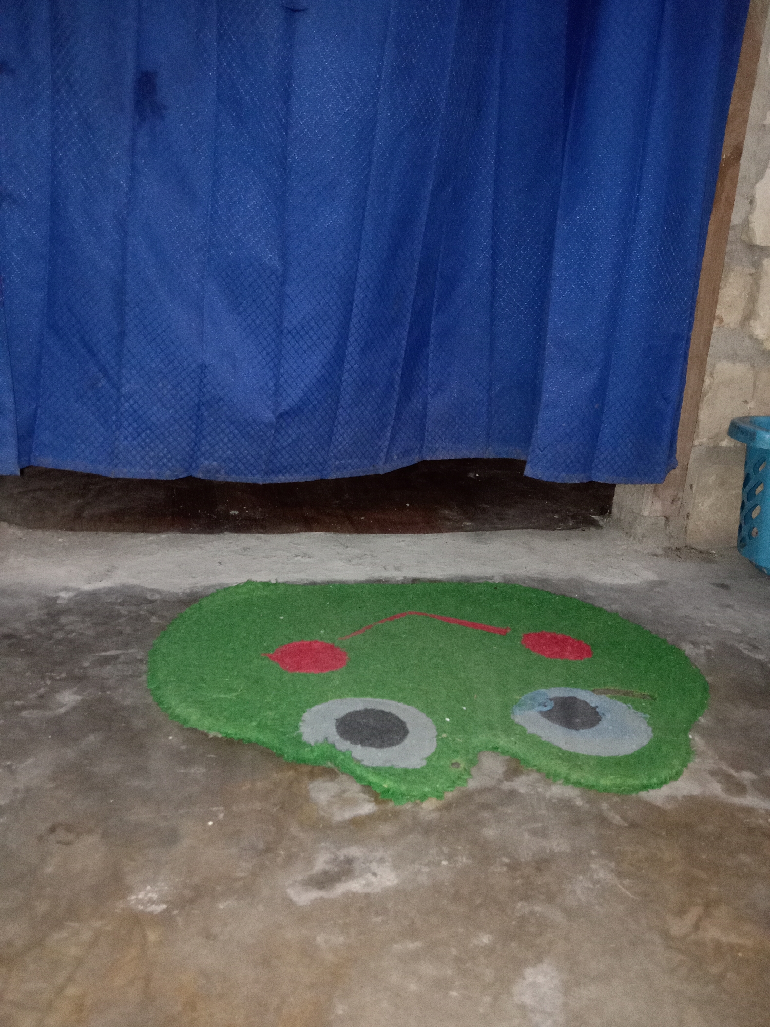 The image shows a green doormat shaped like a frog's face, with red circles for cheeks and black and gray circles for eyes, placed on a concrete floor in front of a blue curtain. The environment appears to be indoors, possibly a room with unfinished walls and a wooden beam on the right side of the frame. A blue plastic basket is visible on the far right. The image does not provide any specific location details within the city of Jumput, Indonesia.