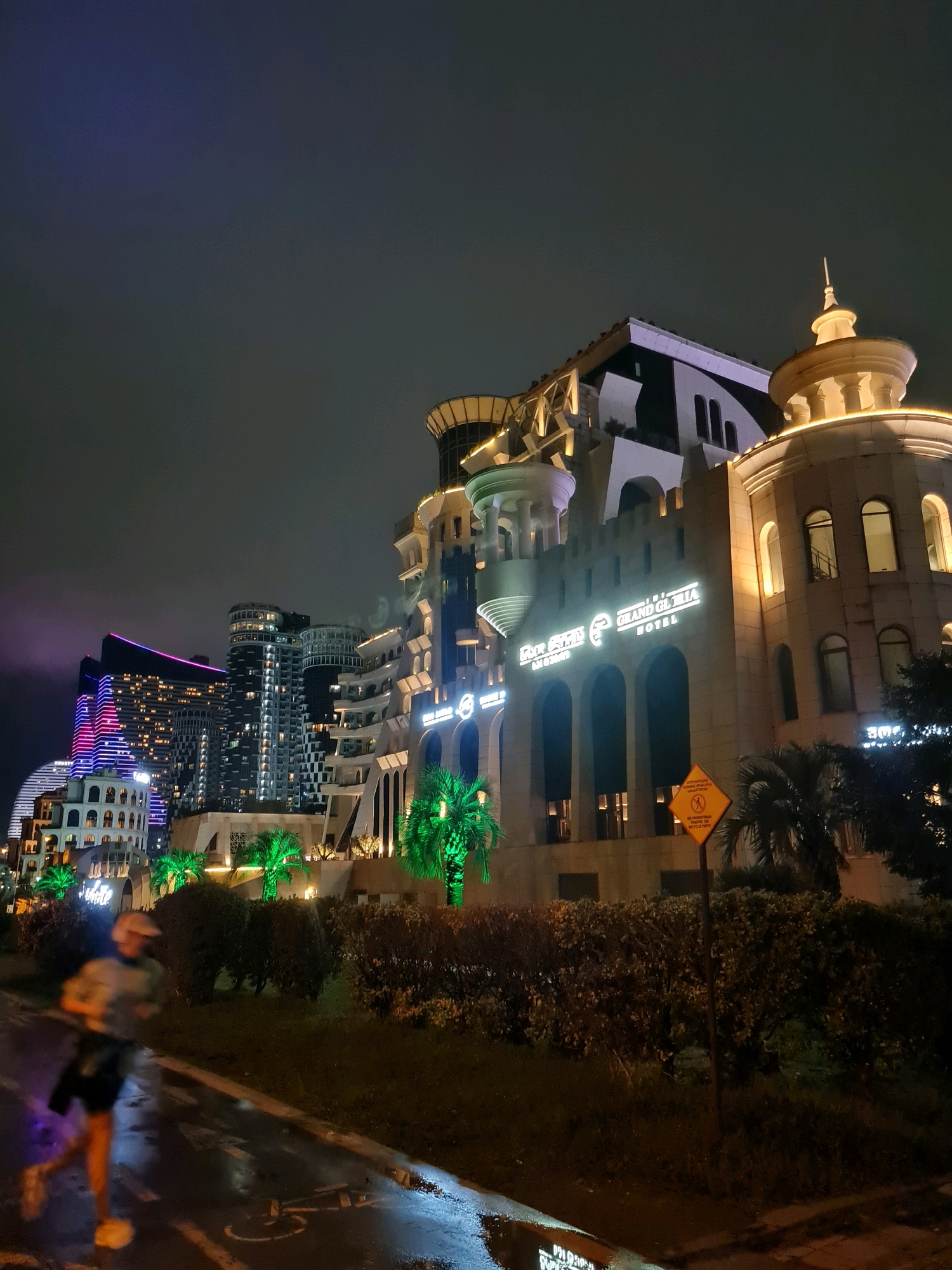 The image depicts a nighttime scene in Batumi, Georgia, specifically along the coastal promenade near the Grand Gloria Hotel. A lone individual, blurred by motion, is captured mid-stride while jogging on a wet asphalt path, presumably after rainfall, indicated by reflections on the surface. The path is marked with painted lines, one of which resembles a bicycle symbol.
Dominating the background are several illuminated modern buildings. To the right, the Grand Gloria Hotel stands out with its ornate, castle-like architecture and glowing yellow lights. To the left, a cluster of contemporary high-rise buildings displays vibrant, multi-colored LED lighting, featuring hues of pink and blue. Tropical palm trees, illuminated with green lights, are scattered throughout the foreground and midground, adding to the ambiance. A yellow diamond-shaped sign with a red border and black symbols, indicating a "No Pedestrian Traffic On Bicycle Path," is visible in front of a lush, manicured hedge. The sky is dark and overcast, typical of an evening or night scene.